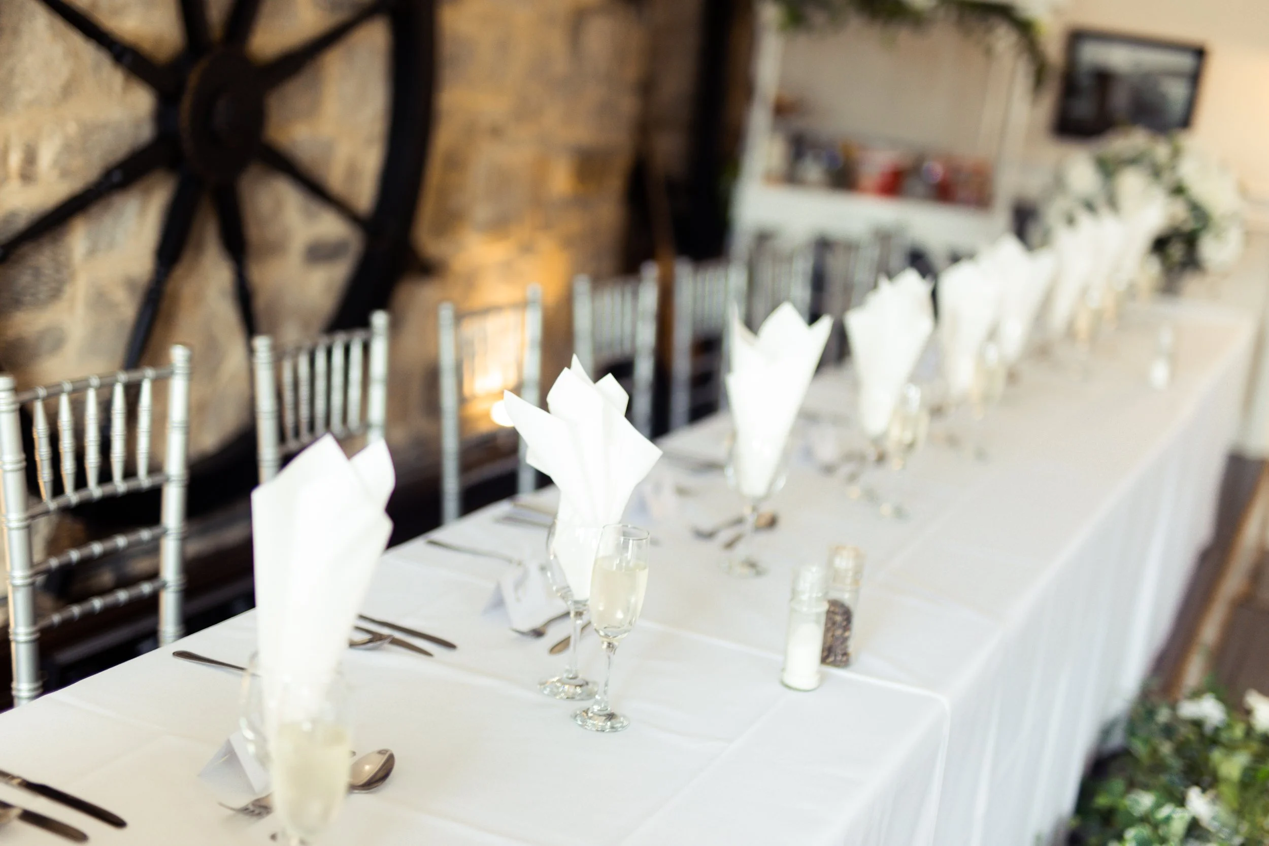Long banquet table set for a formal event with white napkins folded indoors, with chairs, glassware, and salt and pepper shakers, against a rustic brick wall with a large decorative gear.