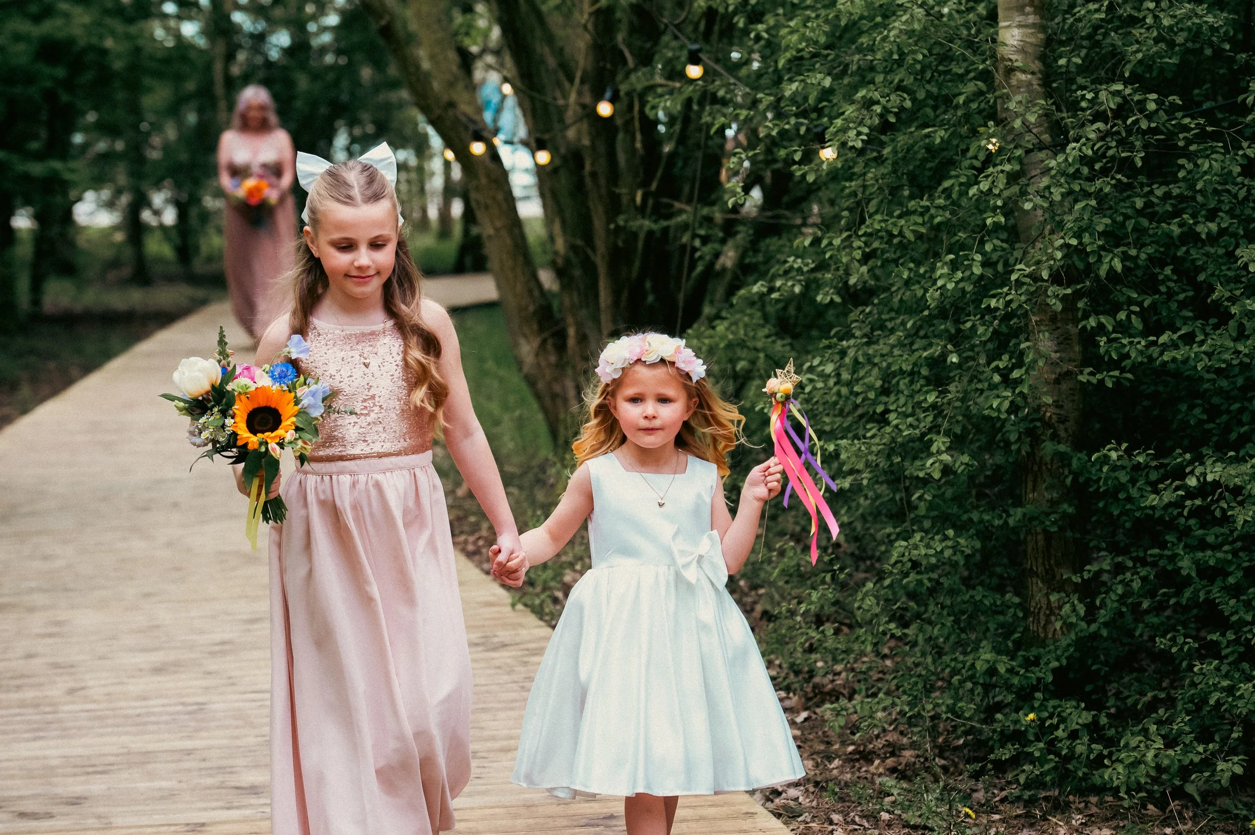 Two young girls, one holding a bouquet of flowers and the other holding colorful ribbons, walk hand in hand on a wooden pathway in a wooded area during daylight. A woman is visible in the background, holding a bouquet, and there are string lights han