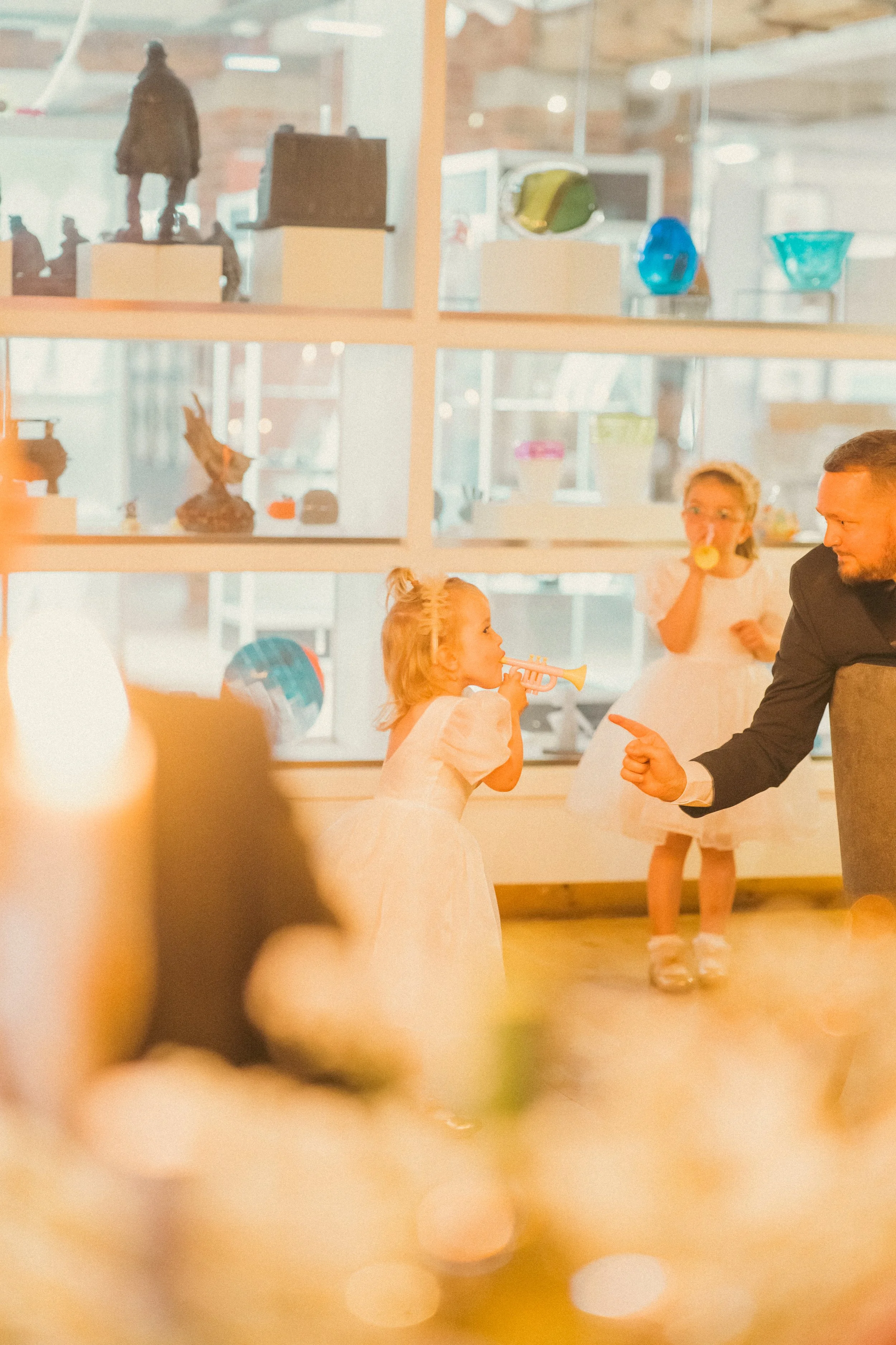Two young girls in white dresses at a celebration, one blowing a trumpet and the other eating a cookie, with a man pointing and interacting with them in a well-lit room decorated for a party.