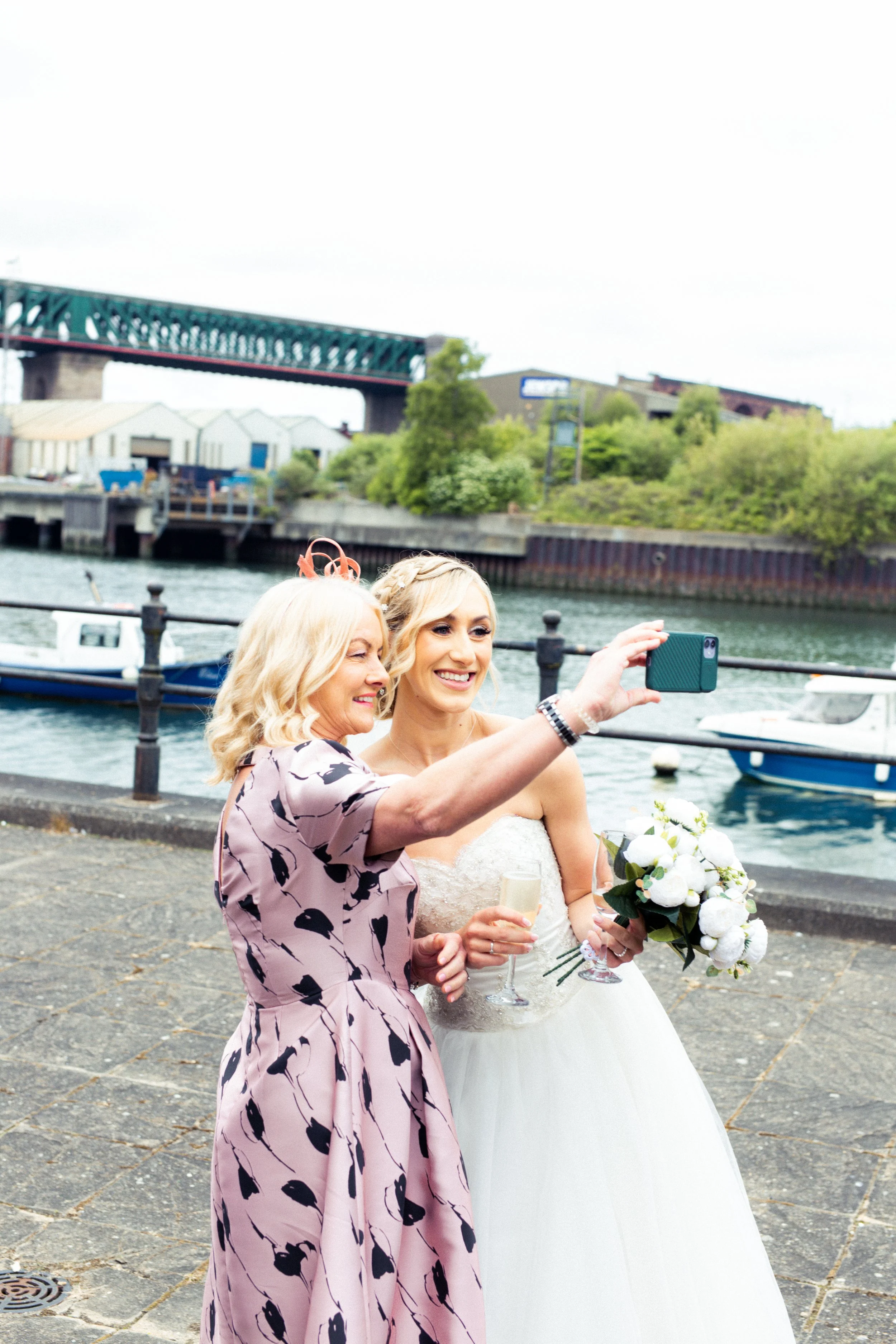 Two women, one bride in a wedding dress holding a bouquet, taking a selfie by a waterfront with boats and a bridge in the background.