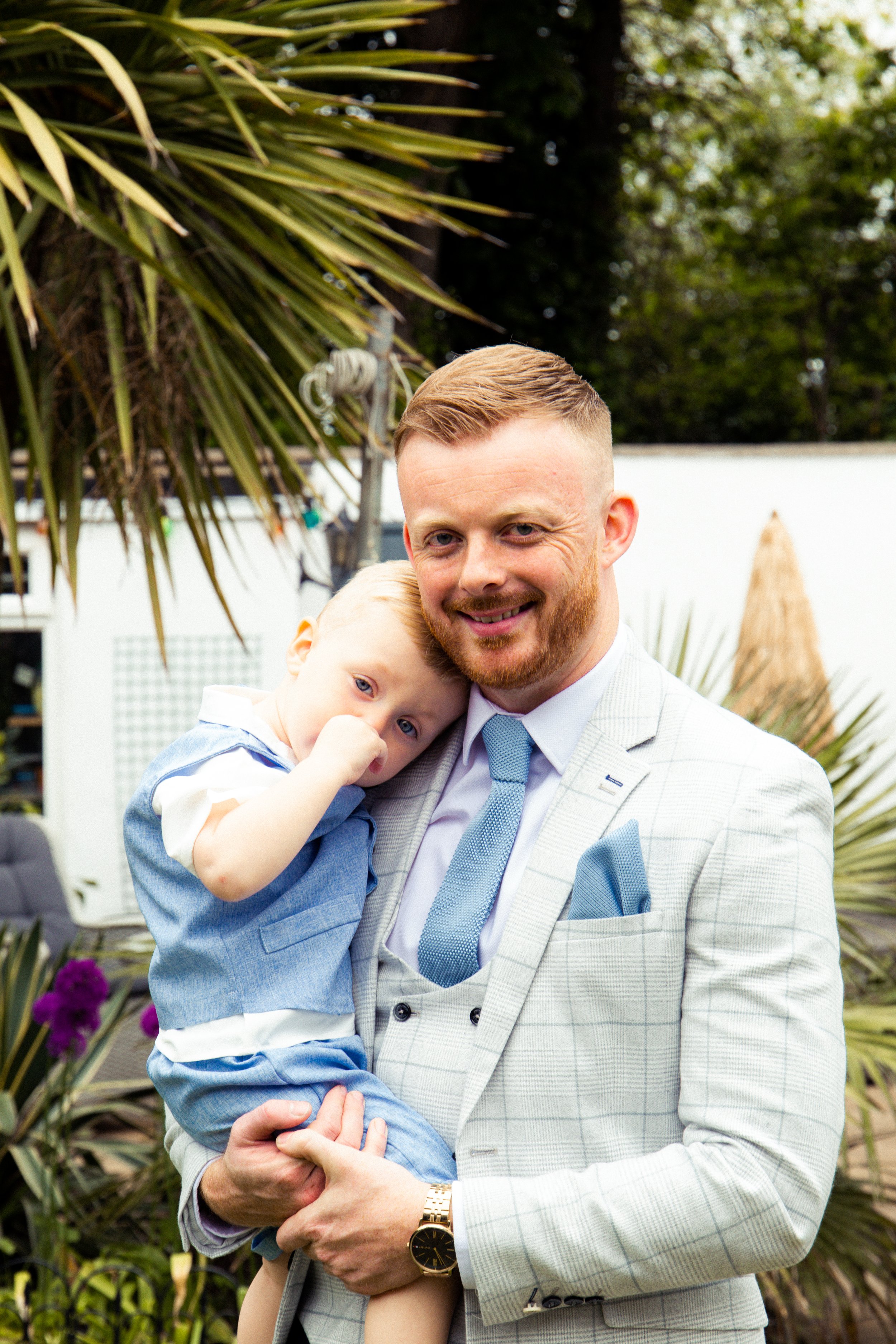 A man in a light-colored suit holding a young child with blond hair, outdoors with greenery in the background.