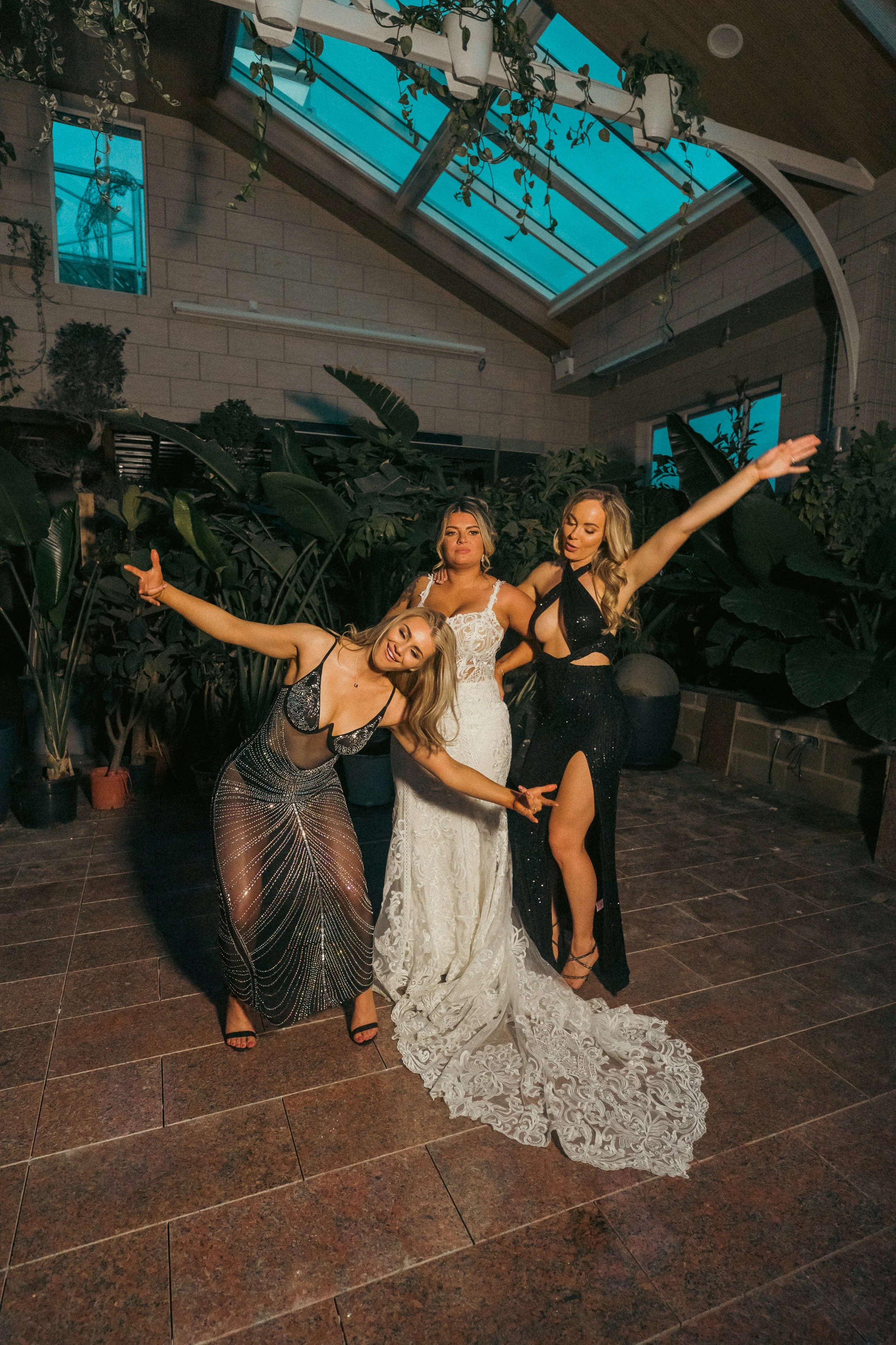 Three women in glamorous dresses posing and dancing indoors with green plants and large windows.