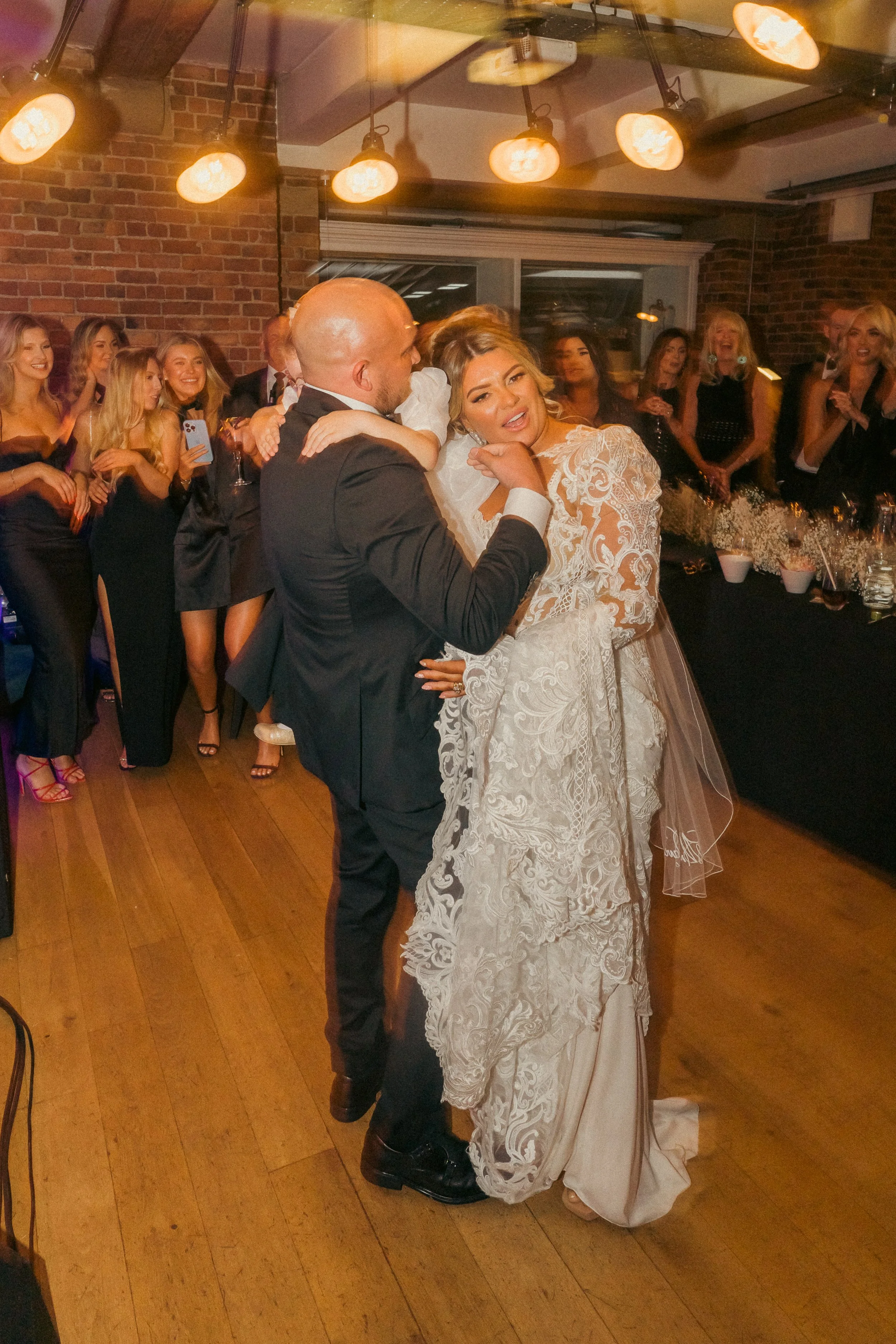 A wedding reception with the bride and groom dancing, surrounded by guests in formal attire, in a brick-walled venue with warm lighting.