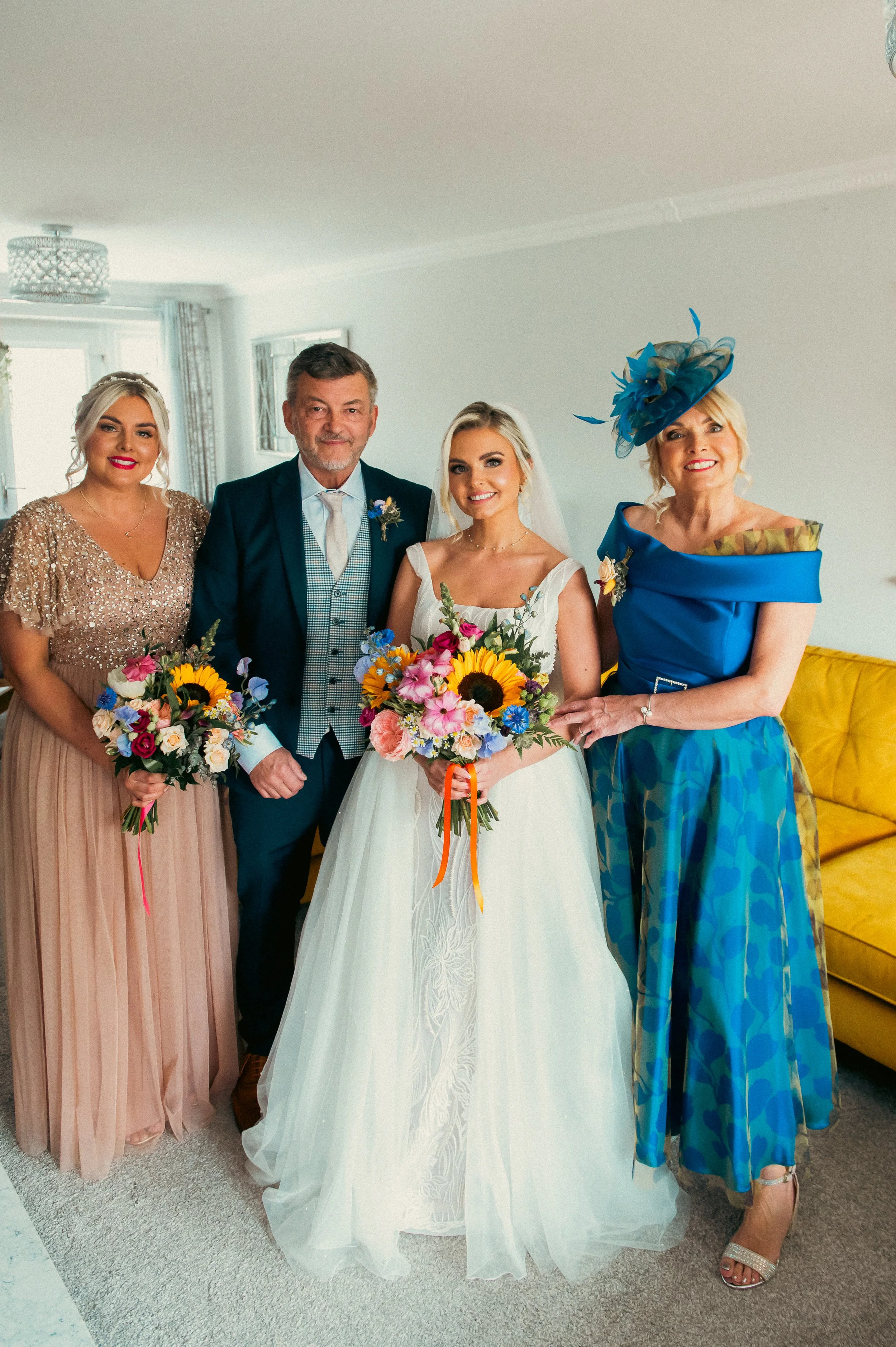 Group of four people, two women, one man, and one girl, dressed in formal attire, standing indoors for a wedding celebration, holding floral bouquets, with a yellow sofa in the background.