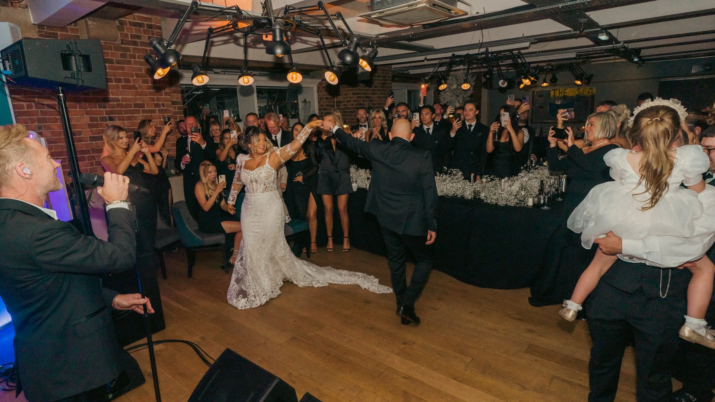 A bride and groom dance together on a dance floor at their wedding reception, with guests watching and taking photos in a dimly lit venue with brick walls.