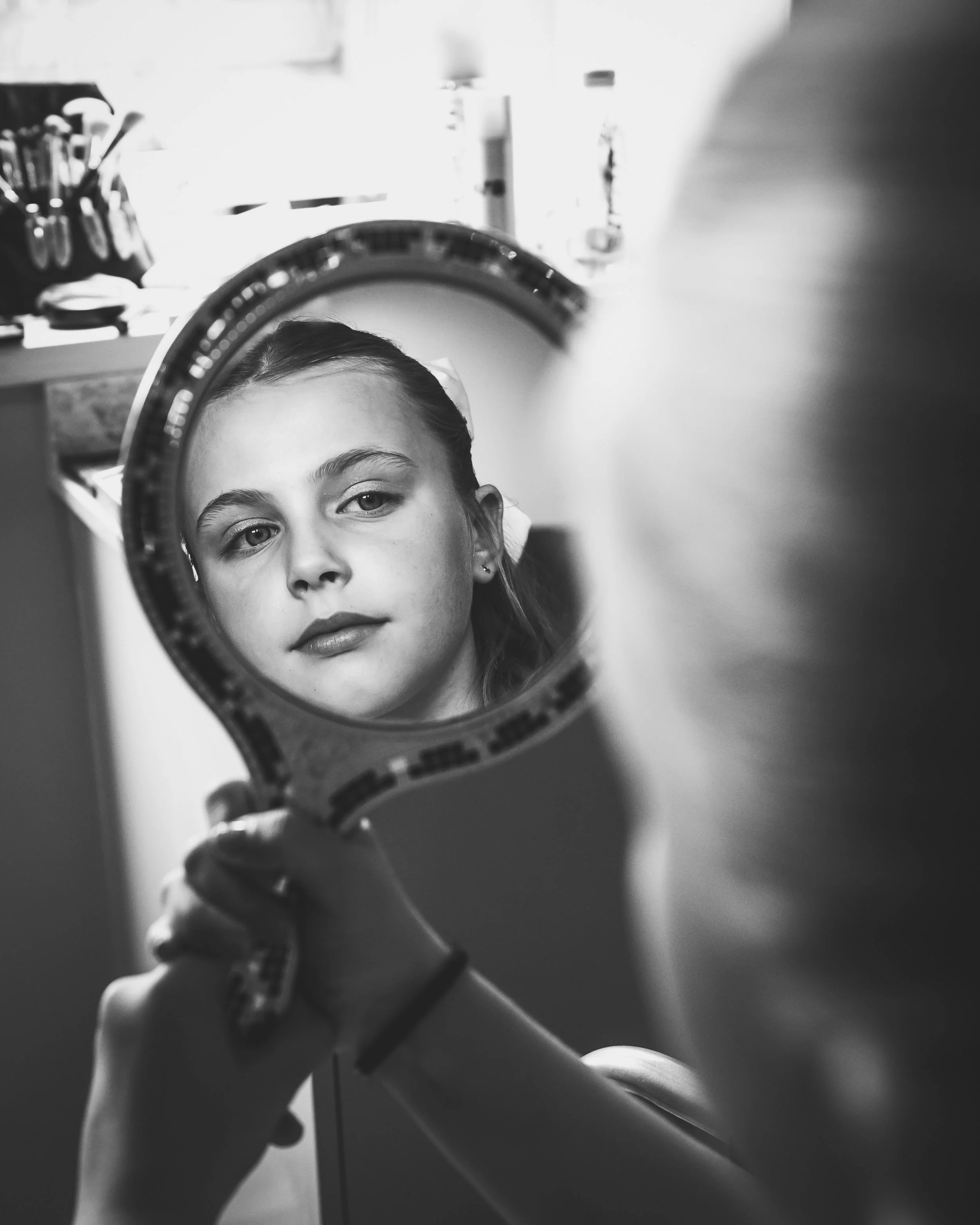 A girl looking into a mirror, her reflection visible in the mirror she is holding, black and white photo.