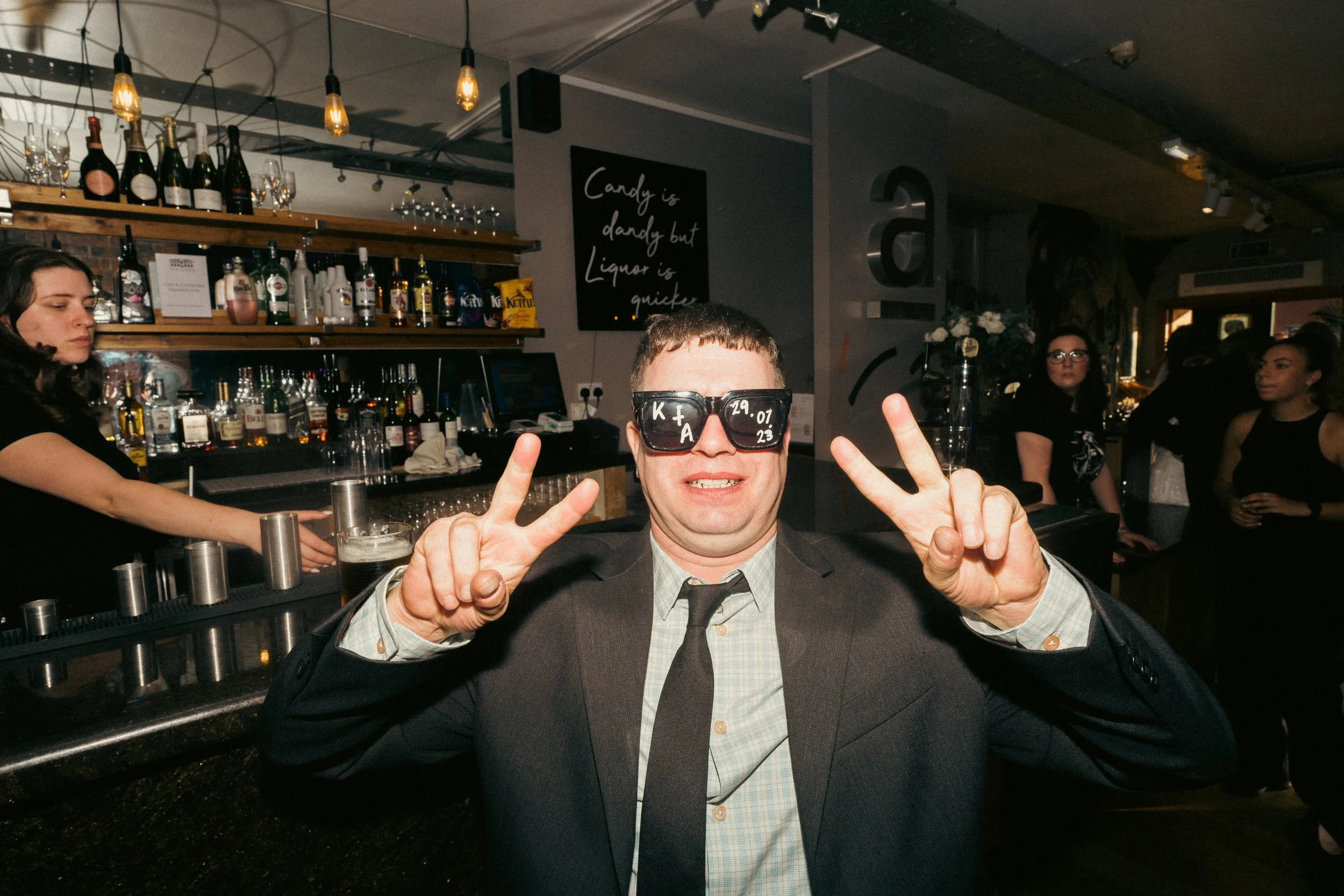 A man in a suit and tie wearing sunglasses with writing on them, making peace signs with both hands, smiling at a bar with liquor bottles and glasses behind him, and other people in the background.