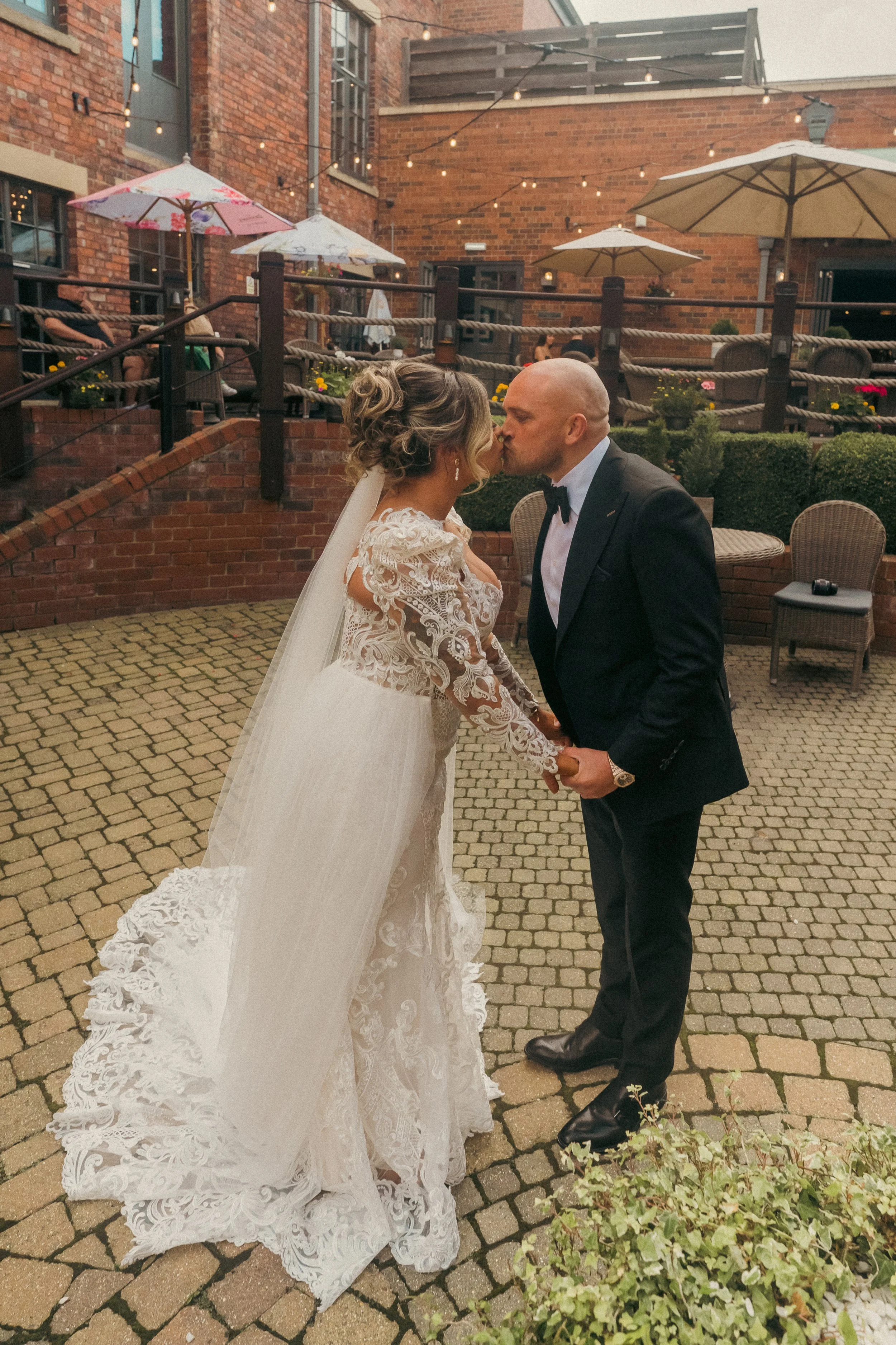 A bride and groom share a kiss during their wedding in an outdoor brick courtyard with string lights and umbrellas.