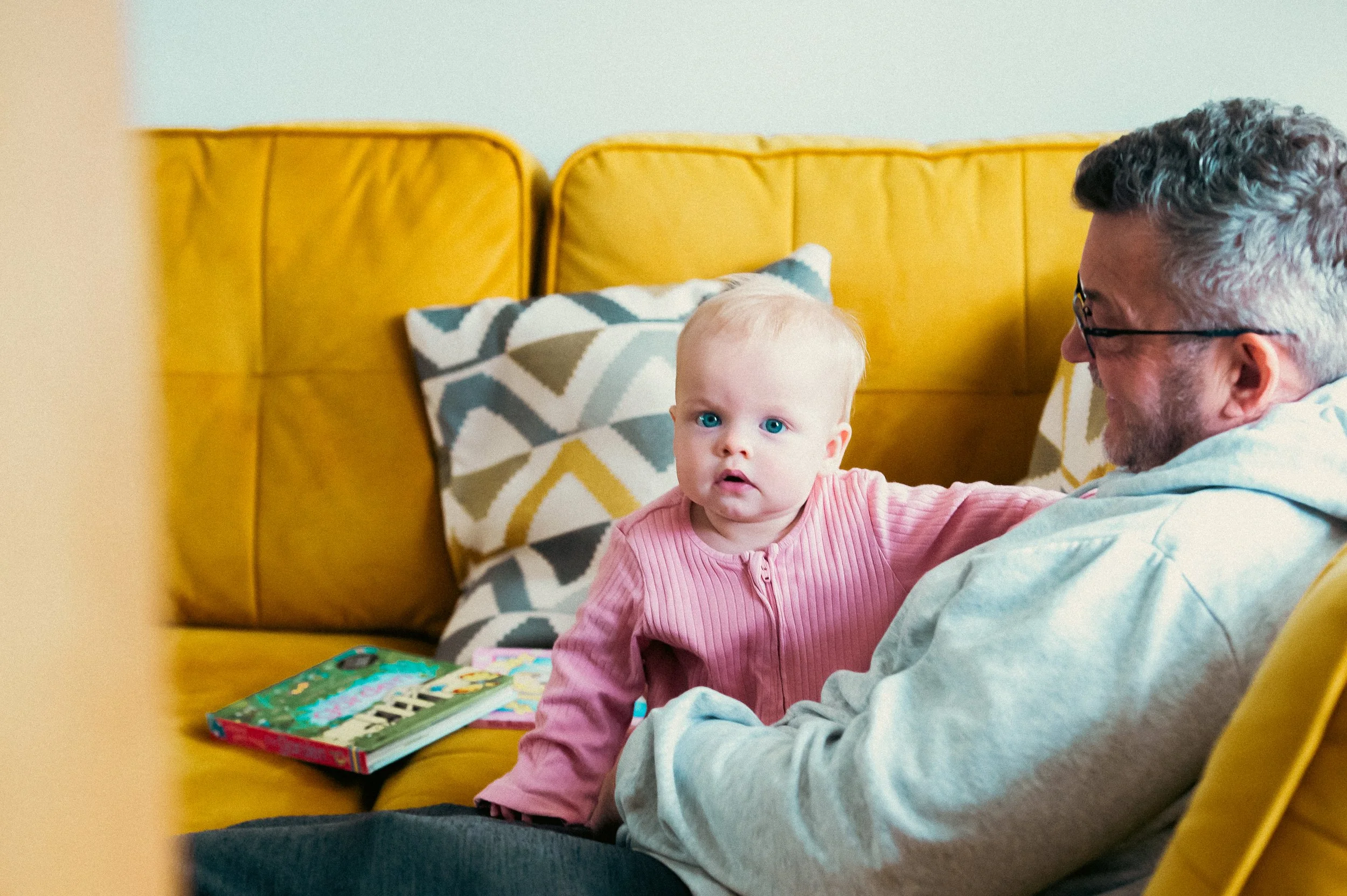 A young child with blonde hair and blue eyes sitting on an older man's lap on a yellow couch, with the child looking surprised or curious. The man is wearing glasses and a gray hoodie. There are colorful books and patterned pillows on the couch.