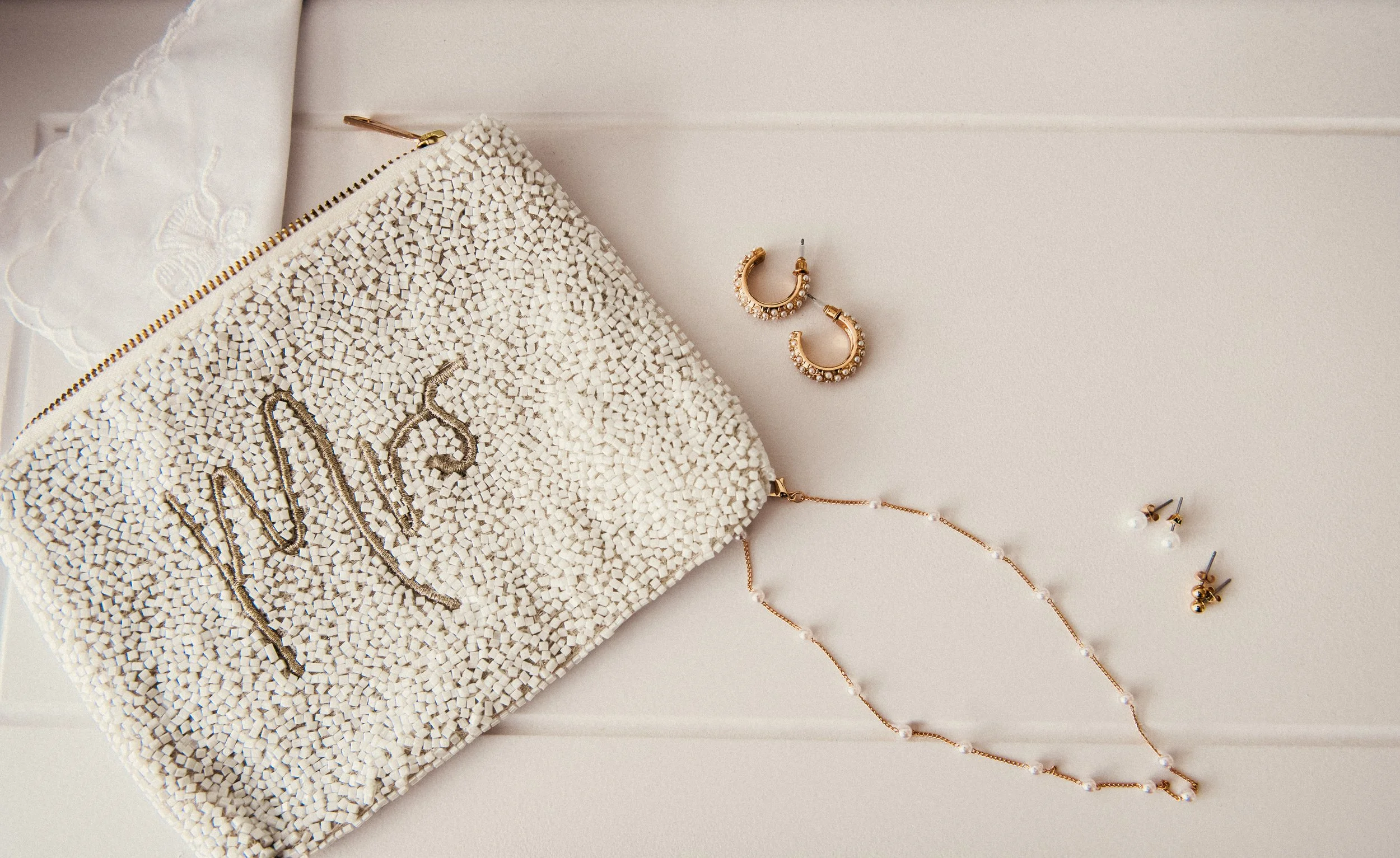 A white beaded clutch purse with the word "Mrs" decorated in rhinestones, placed on a white surface along with pearl and gold earrings, and a pearl necklace.