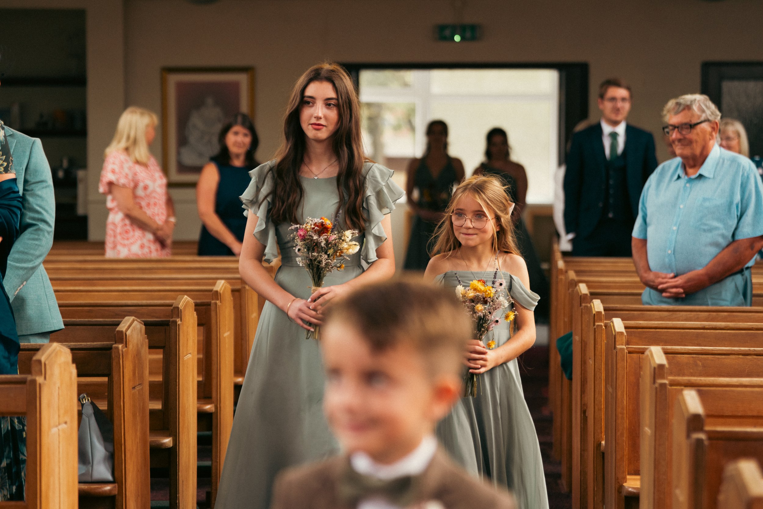 Two young women in gray dresses holding bouquets of flowers walking down an aisle in a church during a wedding ceremony, with guests seated and standing in the background.