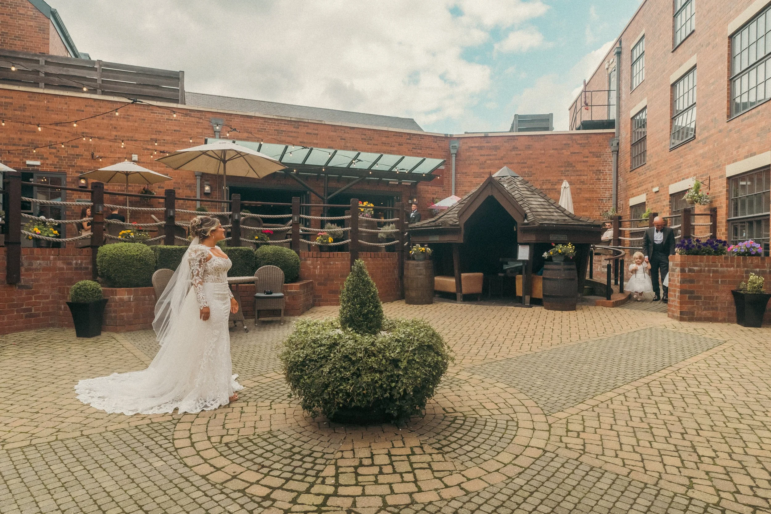 A bride in a white lace wedding dress and veil standing in a courtyard, facing a little girl and an older man who are walking toward her. The courtyard is surrounded by brick buildings, with potted plants, umbrellas, string lights, and a small covere