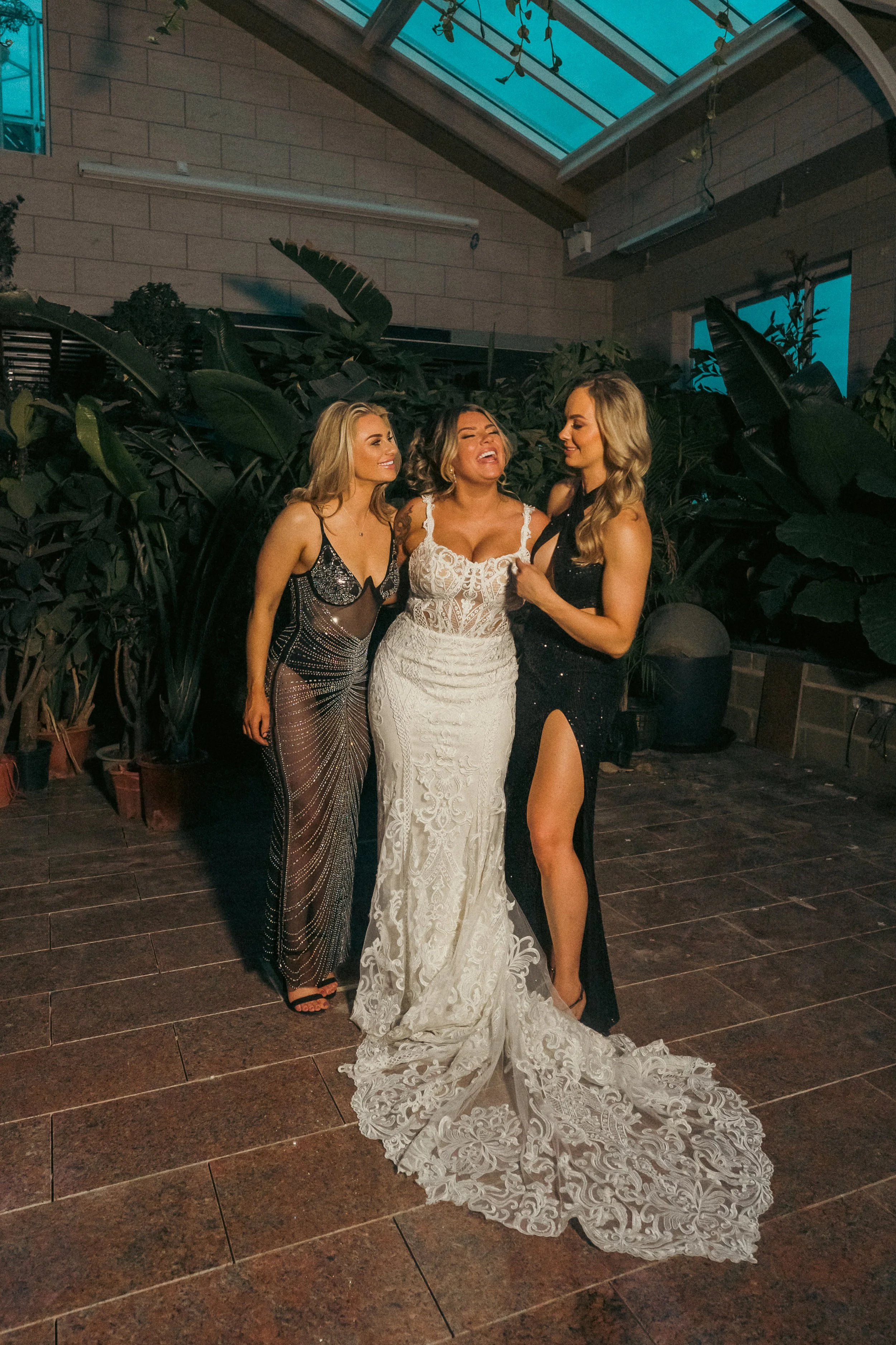 Three women in evening dresses, one in a wedding gown, standing together in an indoor garden or greenhouse with large green plants in the background and a glass ceiling above.