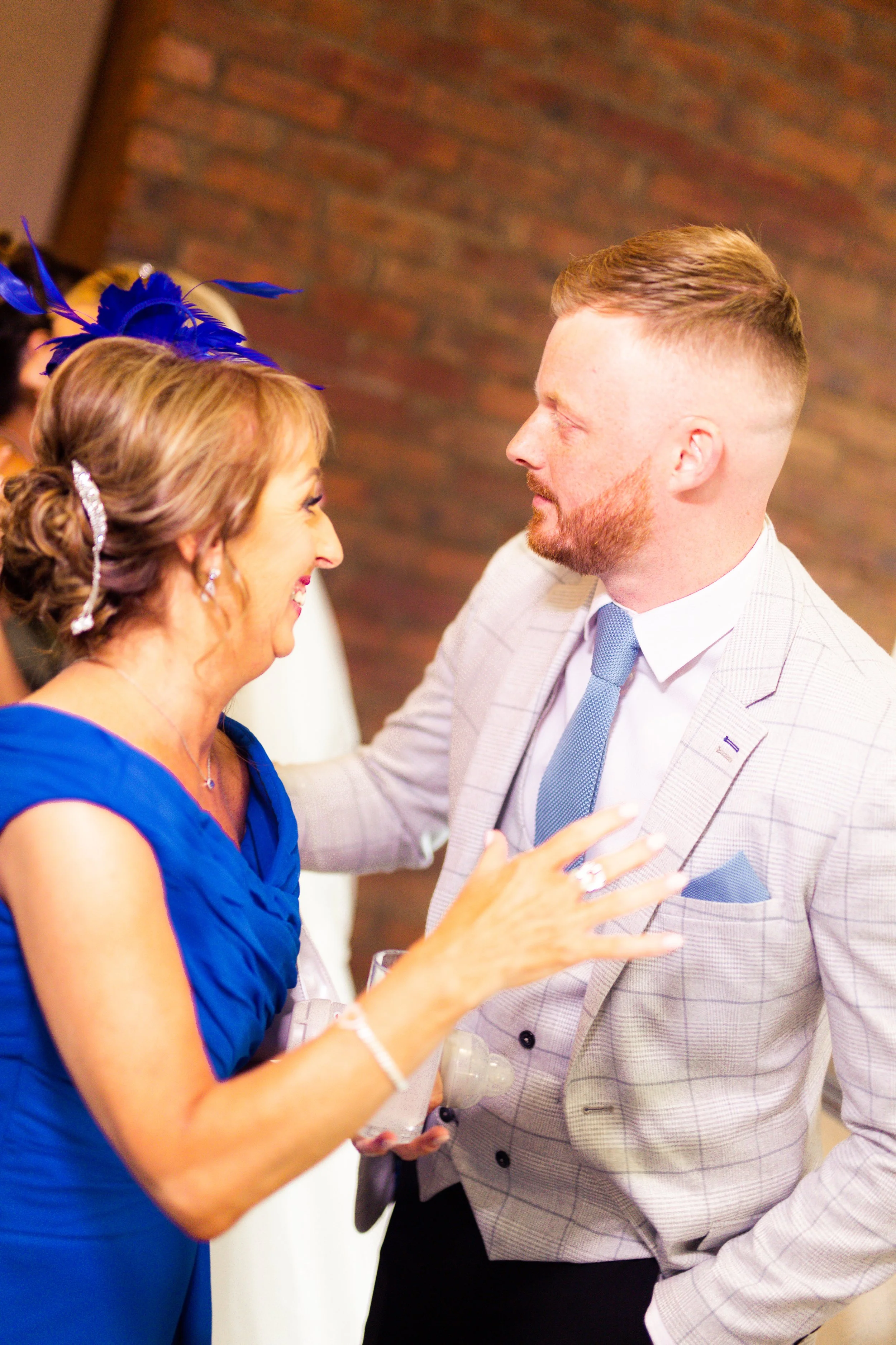 A woman in a blue dress and a man in a light gray suit are smiling and talking closely at a social event. The woman is wearing a blue fascinator and has her hand raised towards the man.