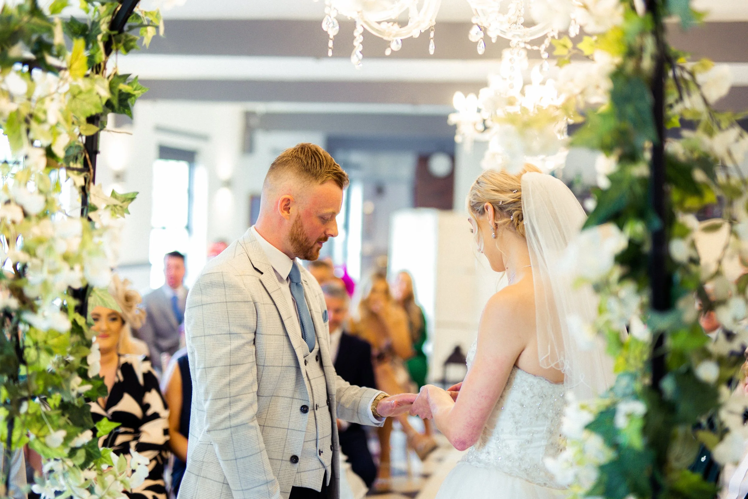 A bride and groom holding hands during their wedding ceremony, surrounded by floral decorations and guests in the background.
