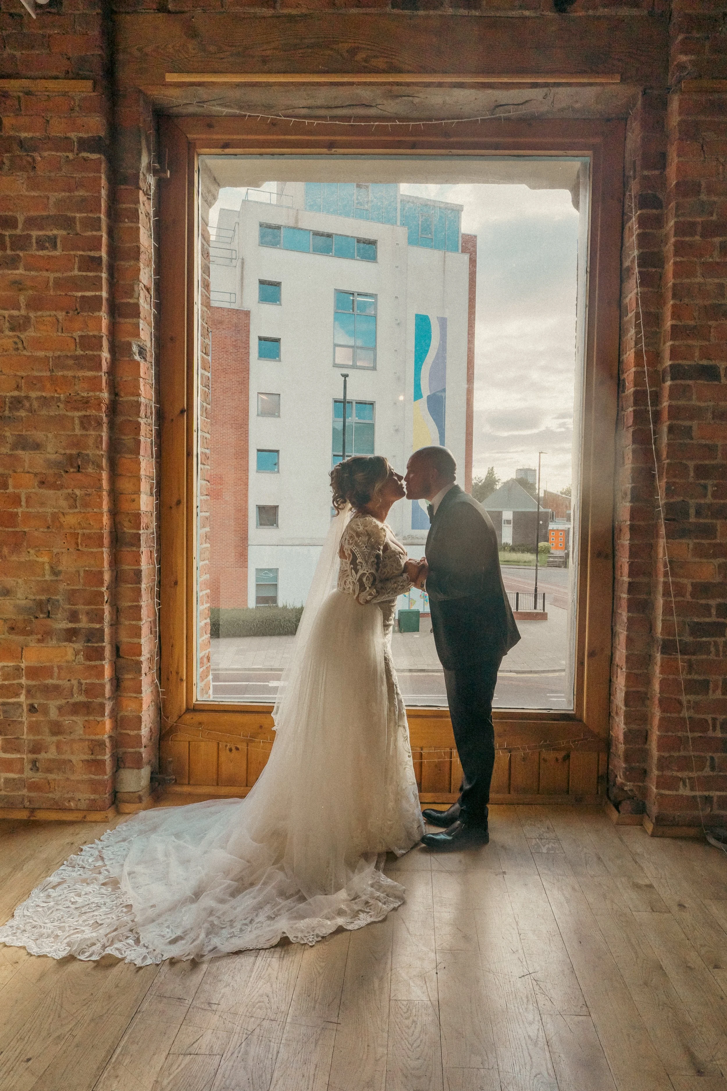 A bride and groom kissing inside a brick building in front of a large window with buildings outside.