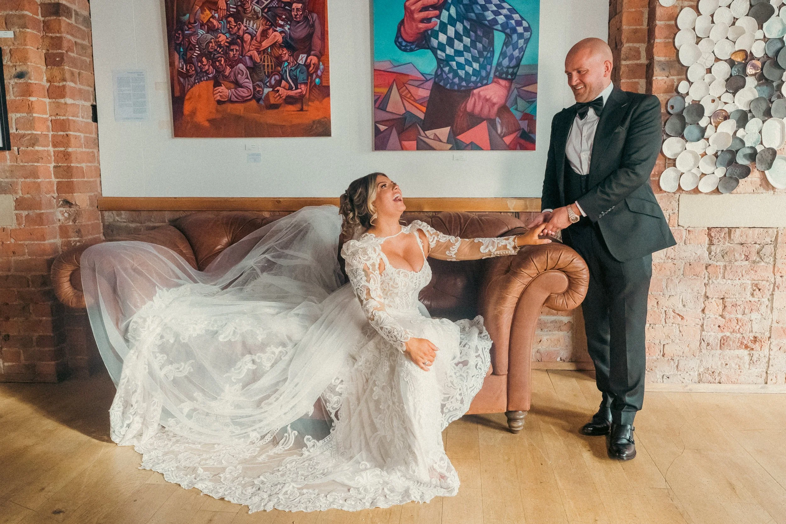 A bride in a white lace wedding gown and veil sitting on a brown leather sofa, laughing and holding hands with a groom in a black tuxedo and bow tie, standing beside her, in an art gallery with colorful paintings and a brick wall background.