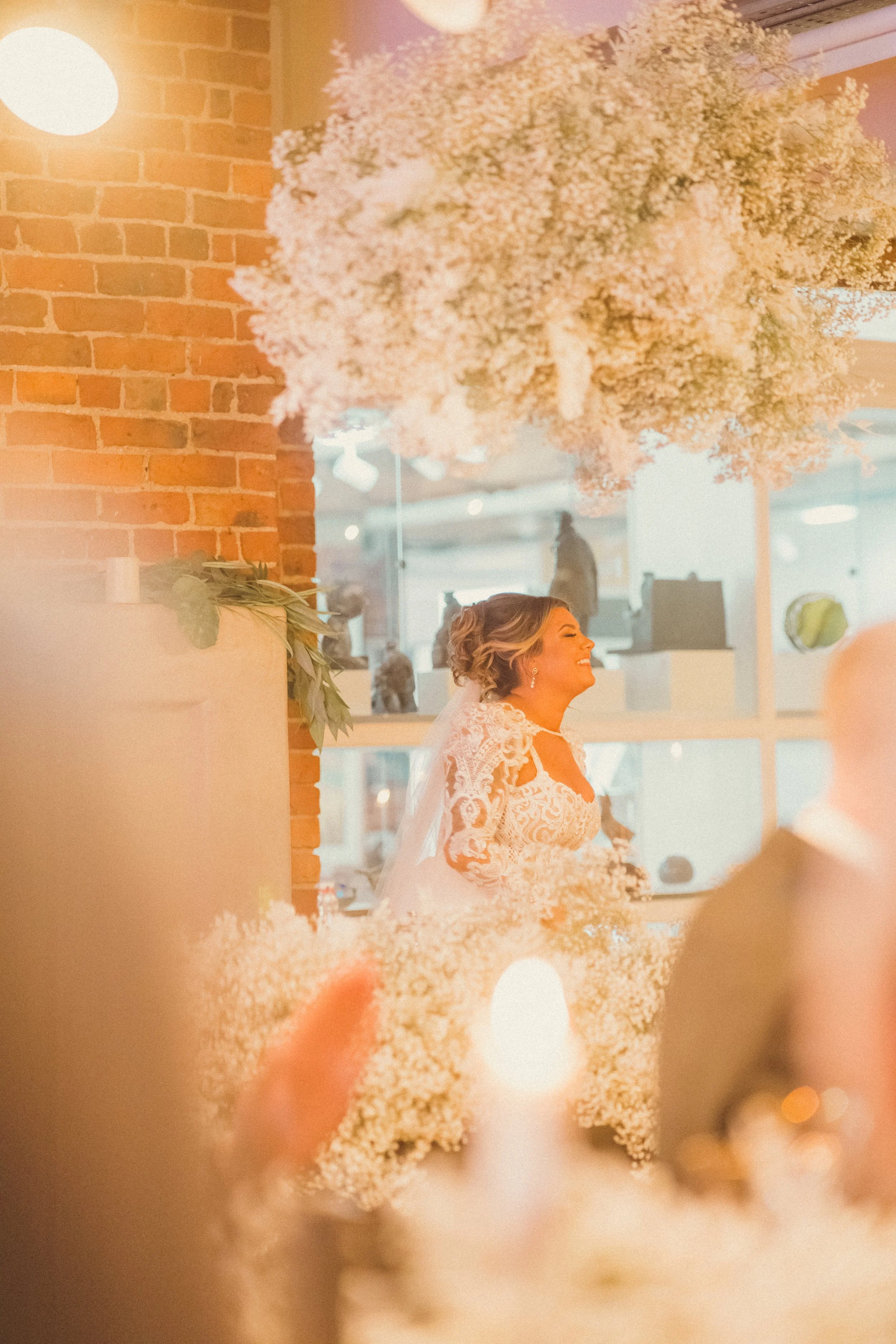 A bride in a white lace wedding dress smiling, standing behind a floral arrangement with baby's breath, inside a venue with brick walls and large windows.