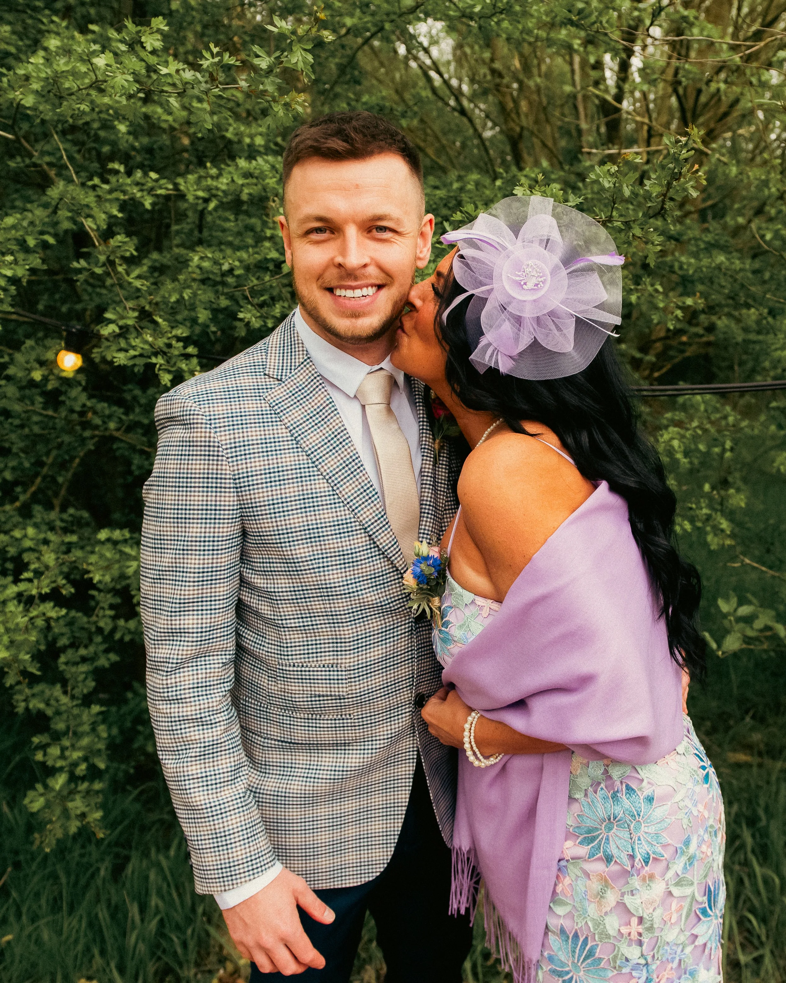 A woman with black hair hugging and kissing a smiling man in a plaid suit outdoors, with trees in the background.