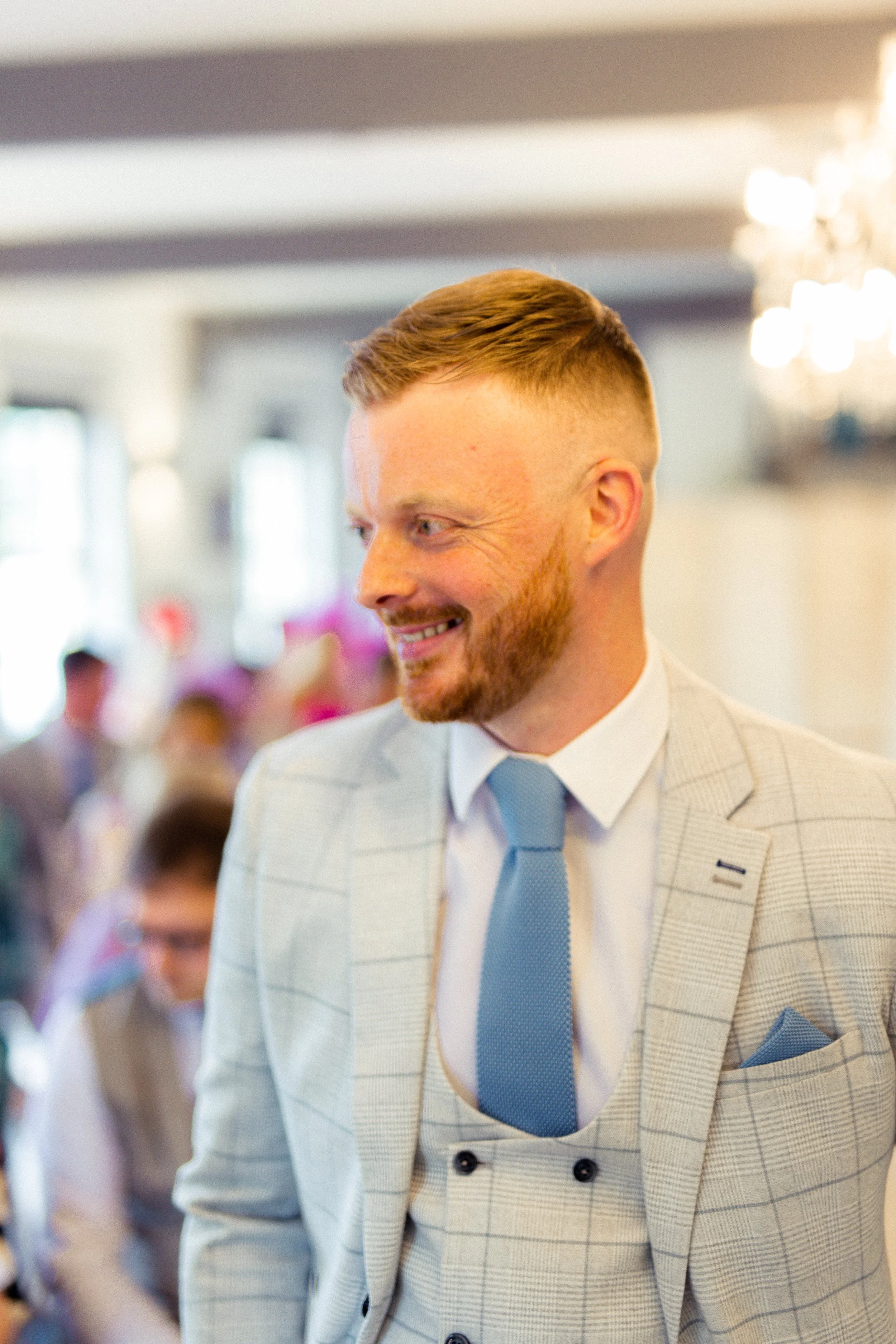 A man dressed in a light-colored plaid suit with a blue tie and pocket square smiling at an event.