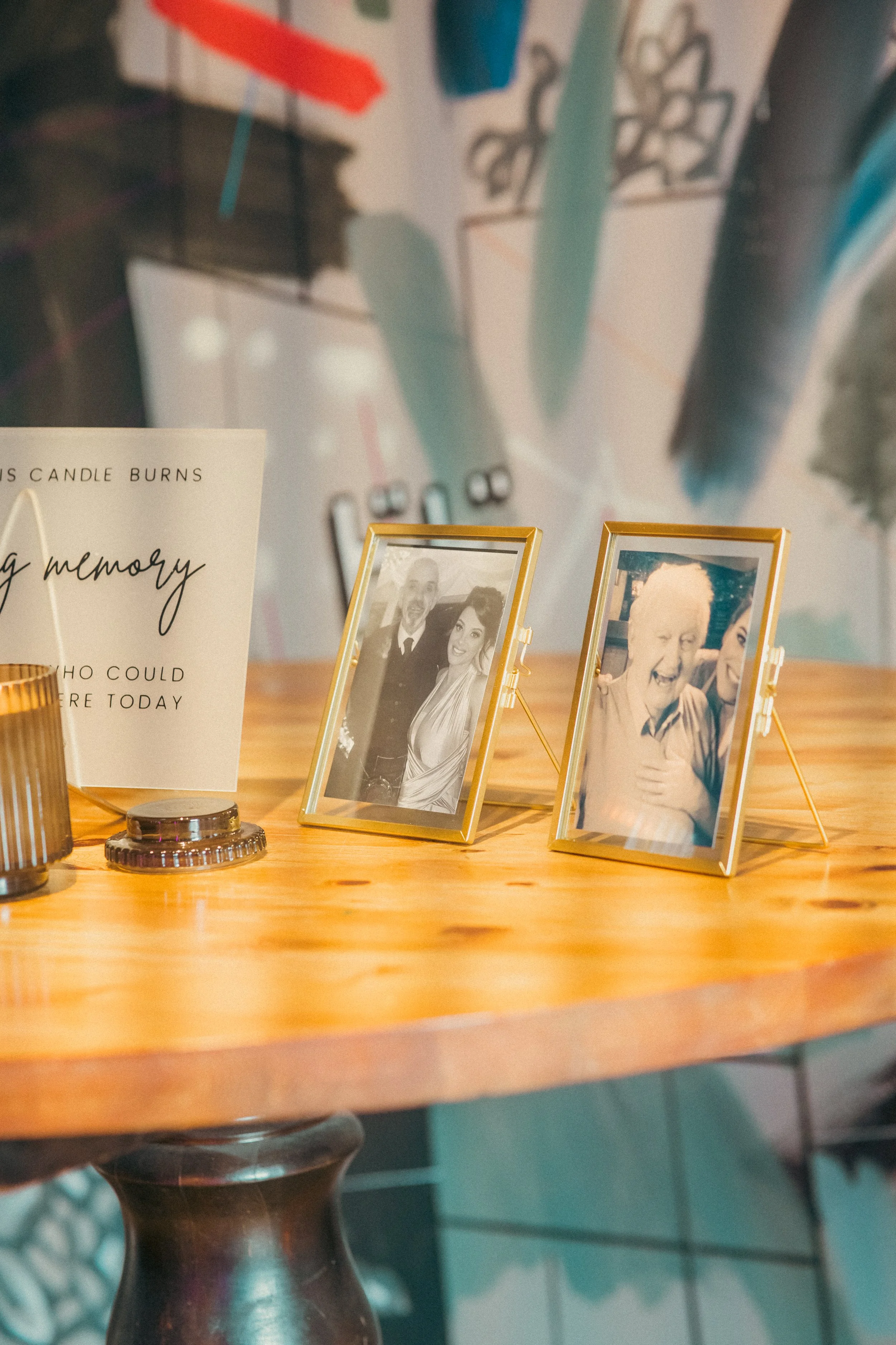 A wooden table with two gold-framed photographs of people smiling, a candle holder, and a card with a heartfelt message.
