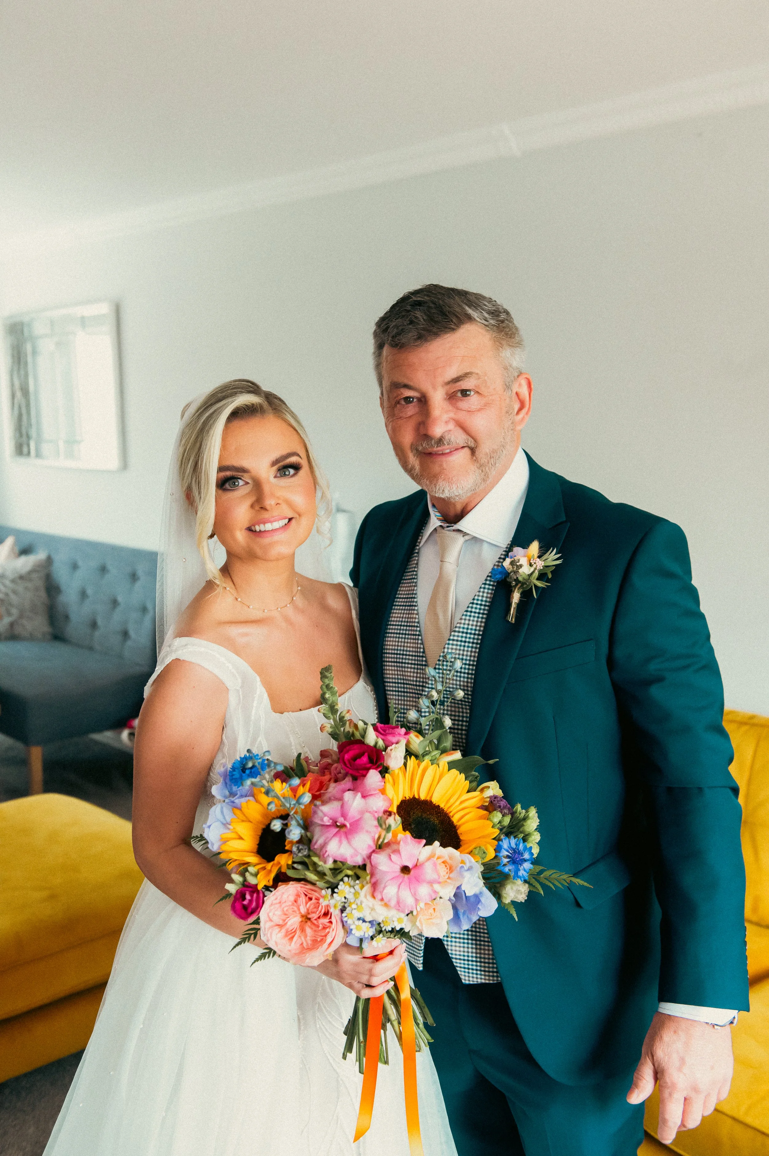 Bride and father smiling indoors, holding a colorful bouquet of flowers.