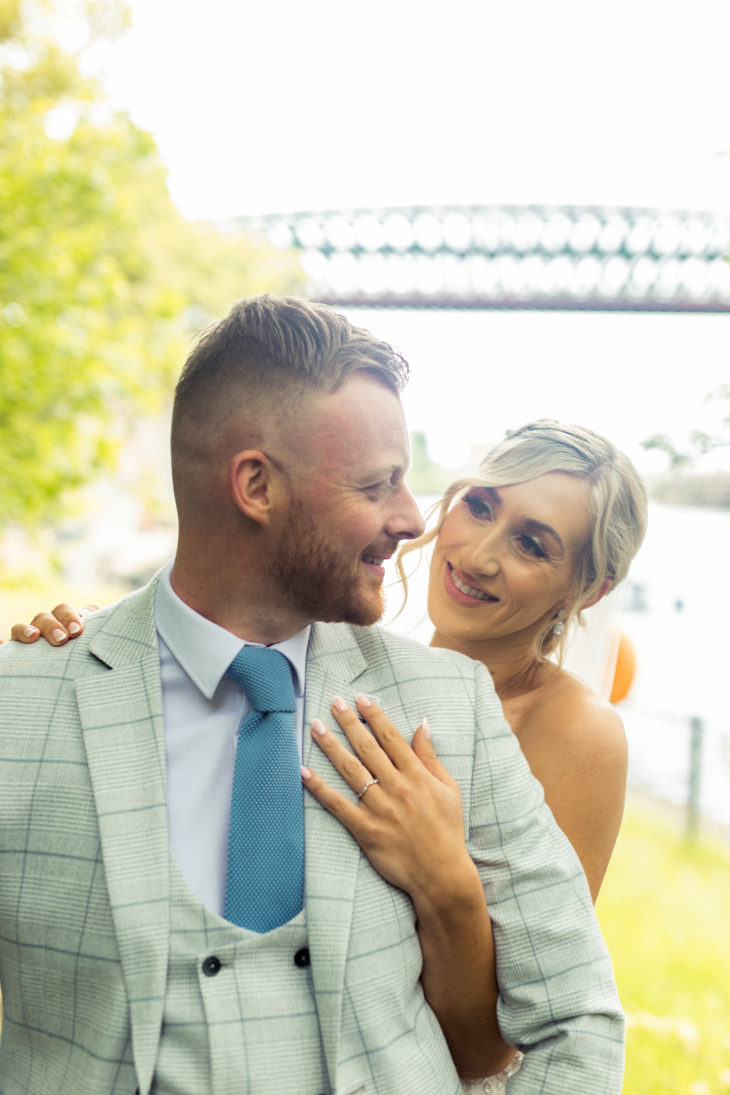 A smiling couple, dressed in wedding attire, embracing outdoors with a bridge in the background.