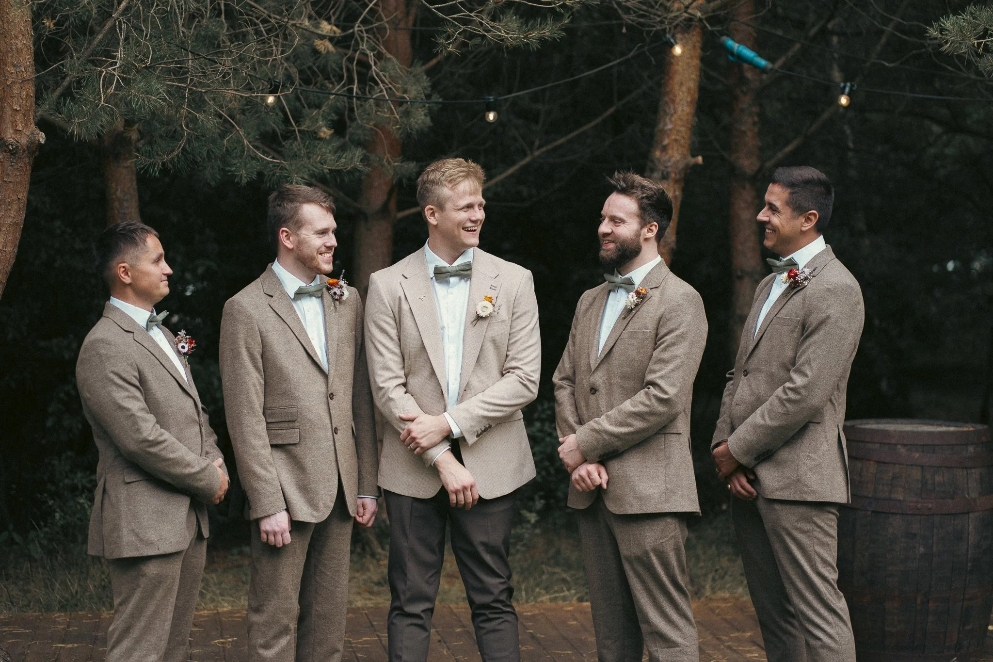 A group of six men dressed in beige suits with bow ties, standing outdoors among trees, smiling and talking during a wedding celebration.