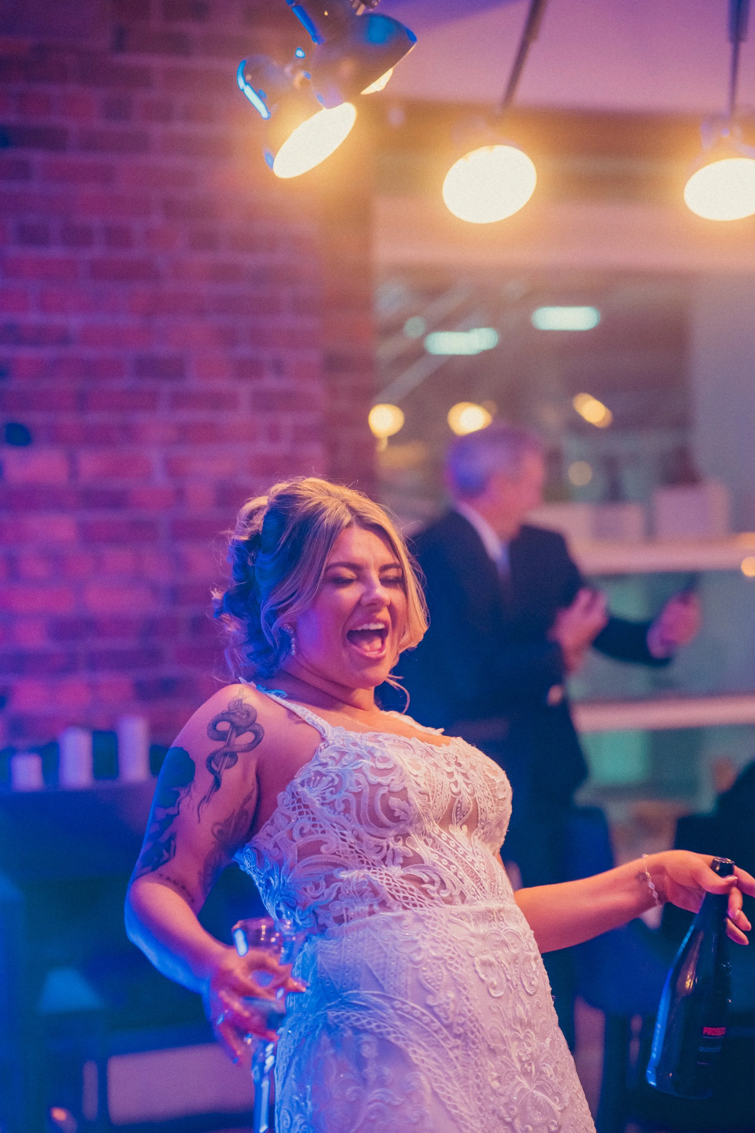 A woman in a white lace dress celebrating at a party, holding a bottle of champagne, with a man in a suit blurred in the background, under colorful lighting with a brick wall behind.