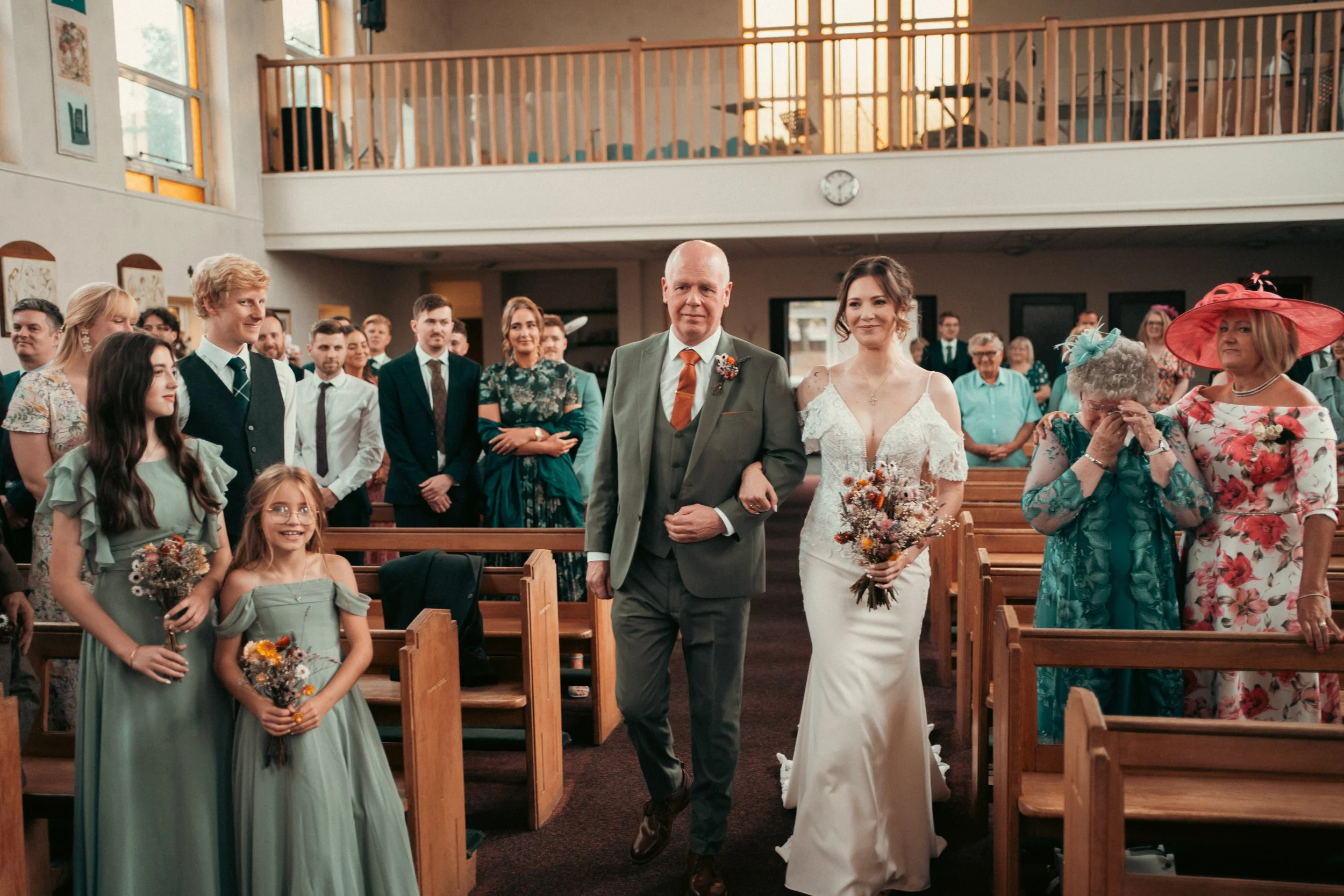 A bride walking down the aisle with her father in a church, surrounded by wedding guests, some standing and some sitting, with floral bouquets in hand.