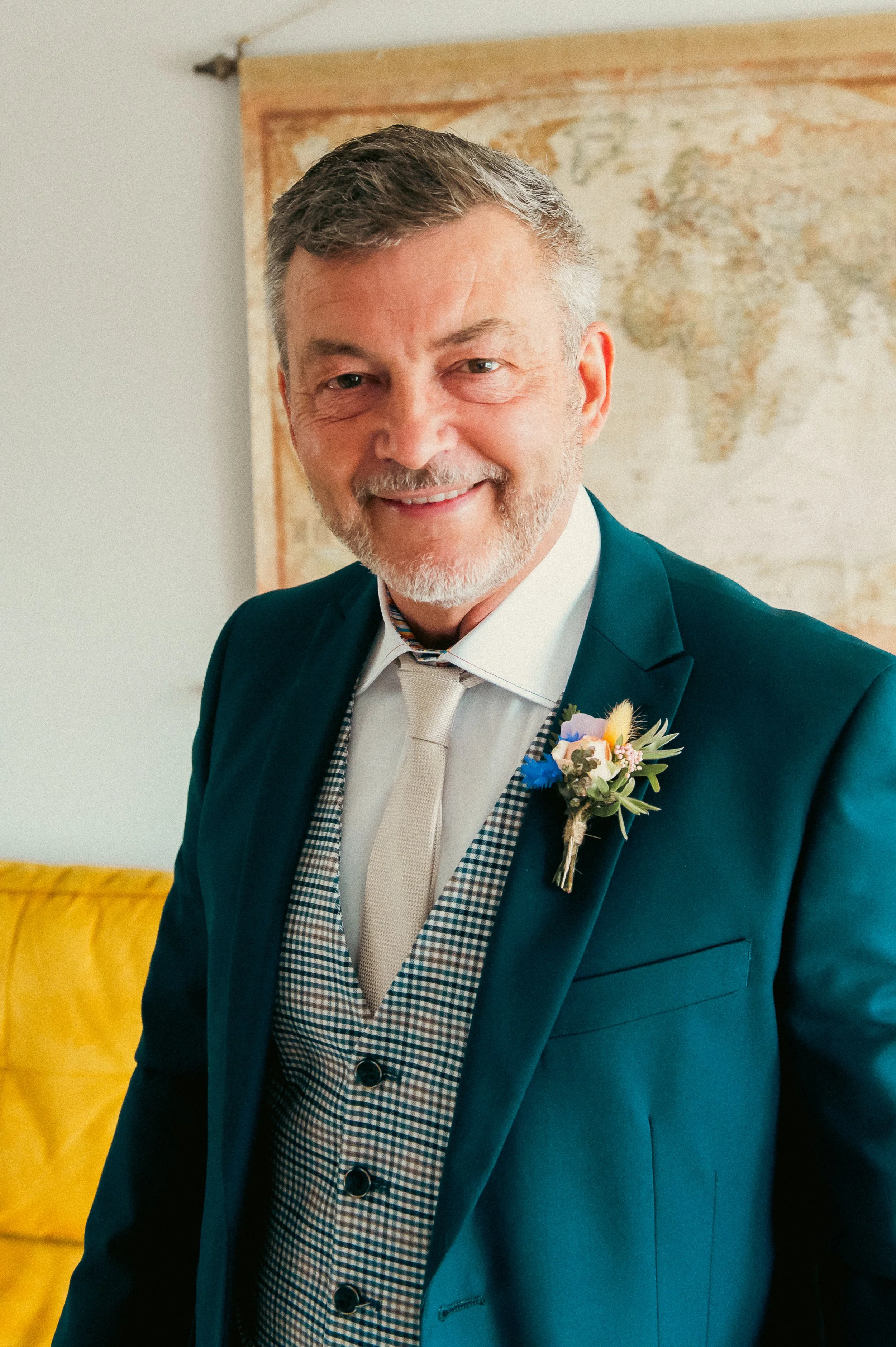 Smiling man in a dark teal suit with a white shirt and light-colored tie, wearing a floral boutonniere, standing in front of a world map.