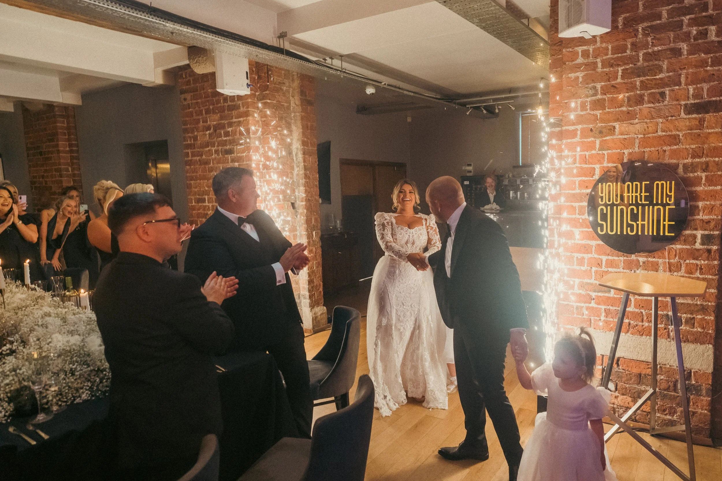 A wedding reception with a bride in a white lace gown smiling, a groom in a black tuxedo, and guests clapping. A young girl in a white dress is holding the groom's hand. The setting features brick walls, string lights, and a sign that reads 'You Are 