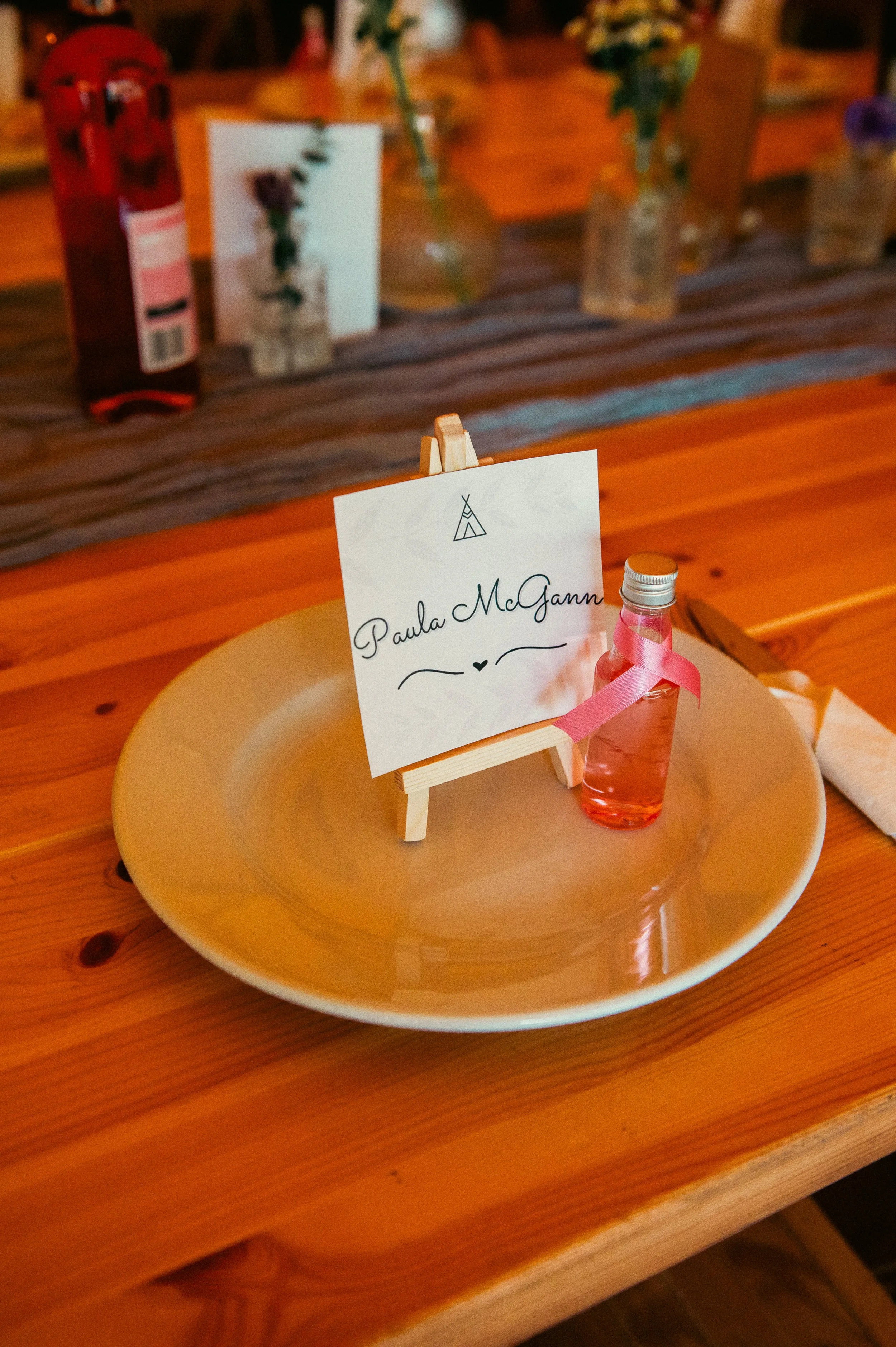 A white plate holds a small glass water bottle with a pink ribbon. An easel displays a sign with the name "Paula McGann" and a small tent icon, indicating a place setting at an event.
