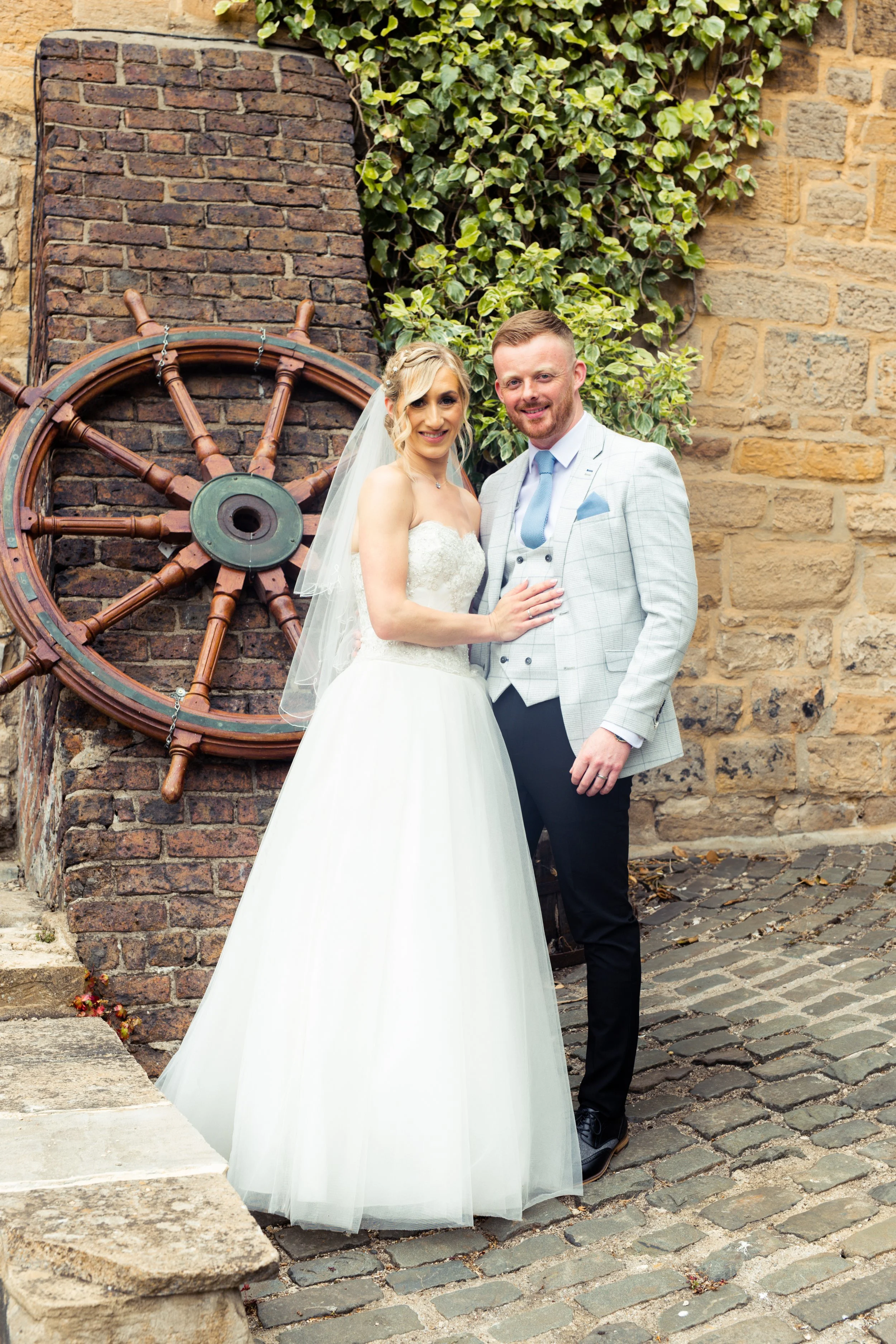 A bride and groom in wedding attire standing close together, smiling, in front of a brick wall with a ship's wheel and green ivy.
