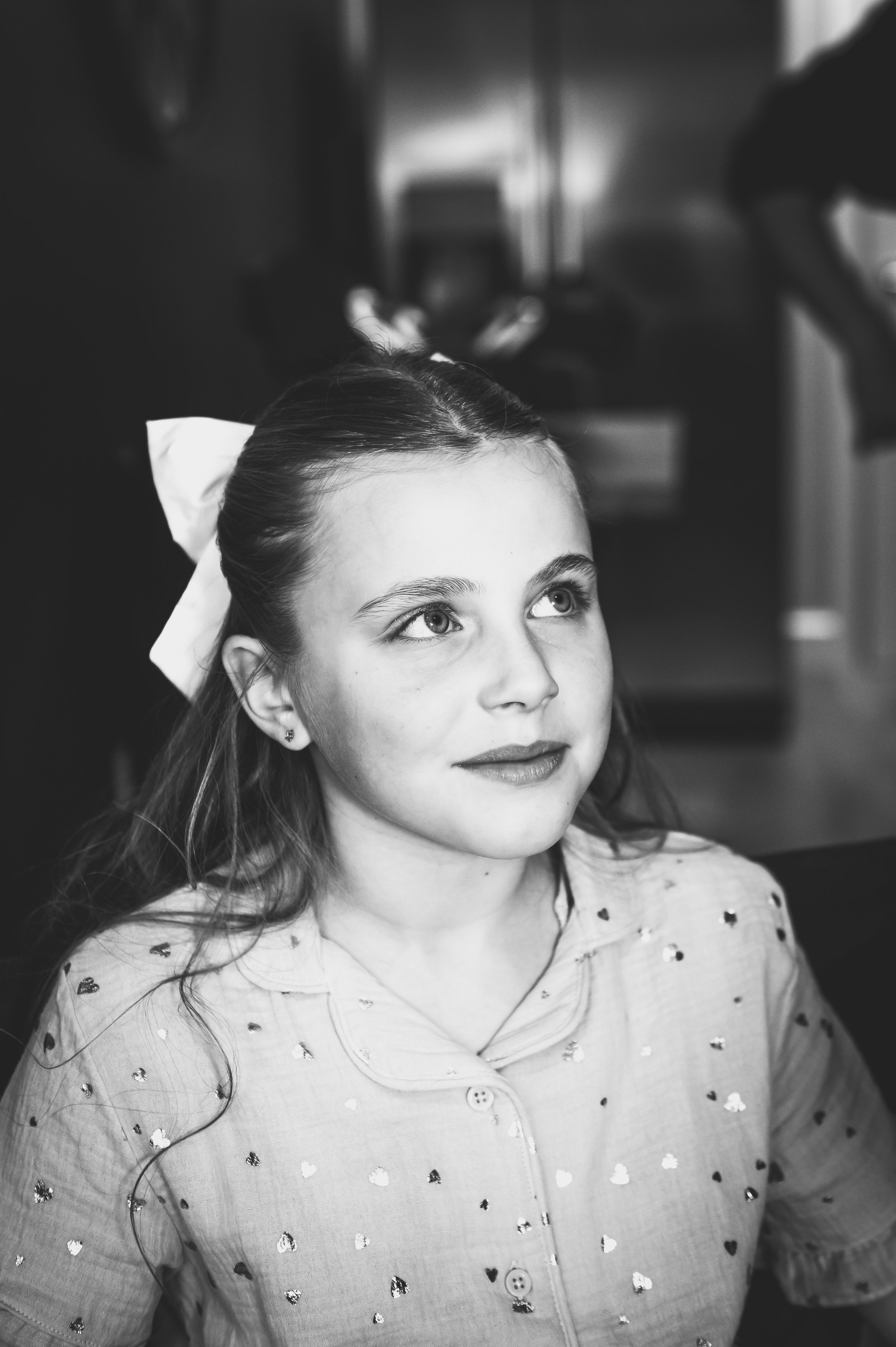 Black and white photo of a young girl with long hair and a white bow, looking upward with a slight smile, wearing a heart-embellished shirt.