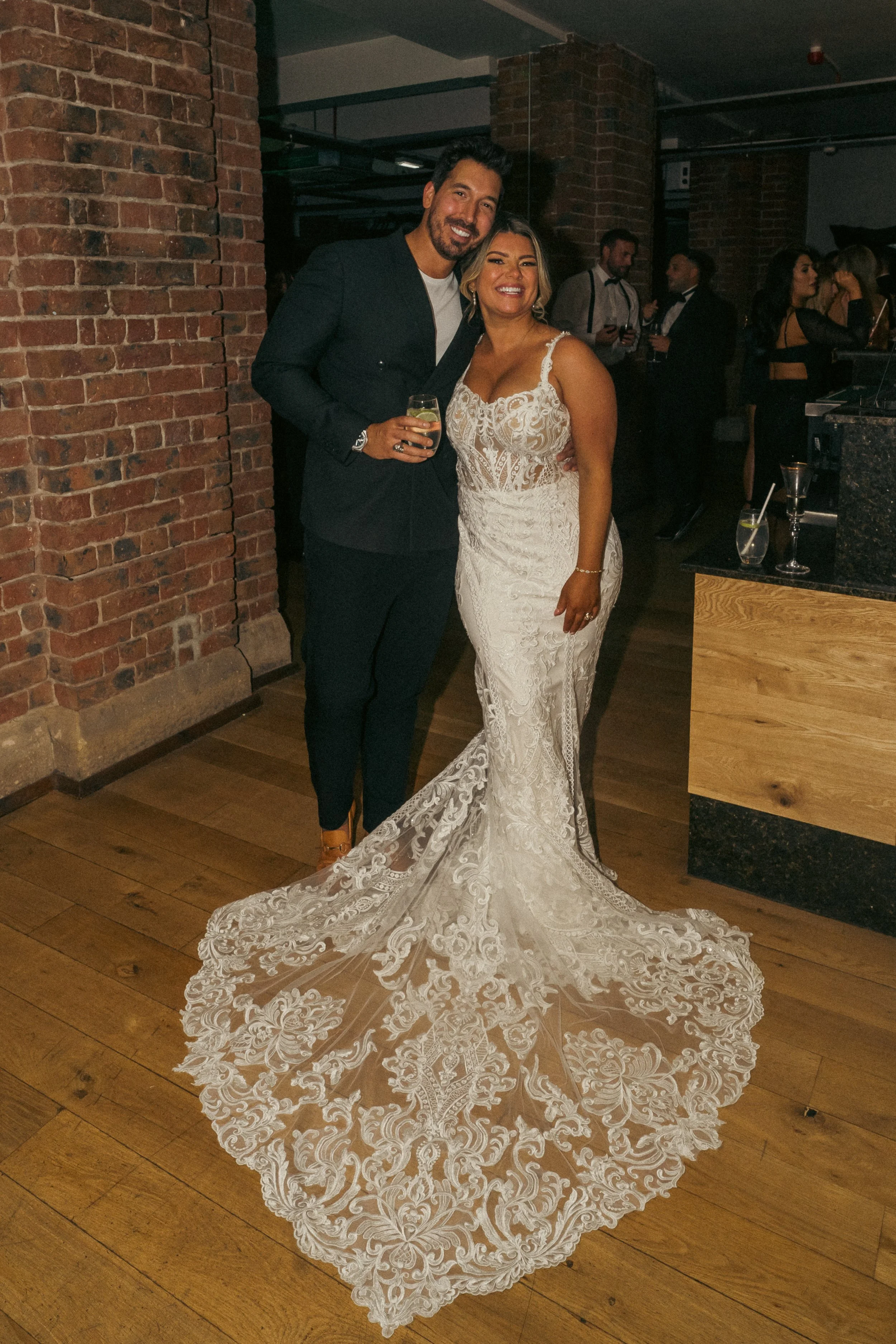 A newlywed couple at a wedding reception, standing together and smiling for the camera. The bride is wearing an elegant lace wedding gown with intricate embroidery and a long train. The groom is dressed in a black suit with a white shirt, holding a w