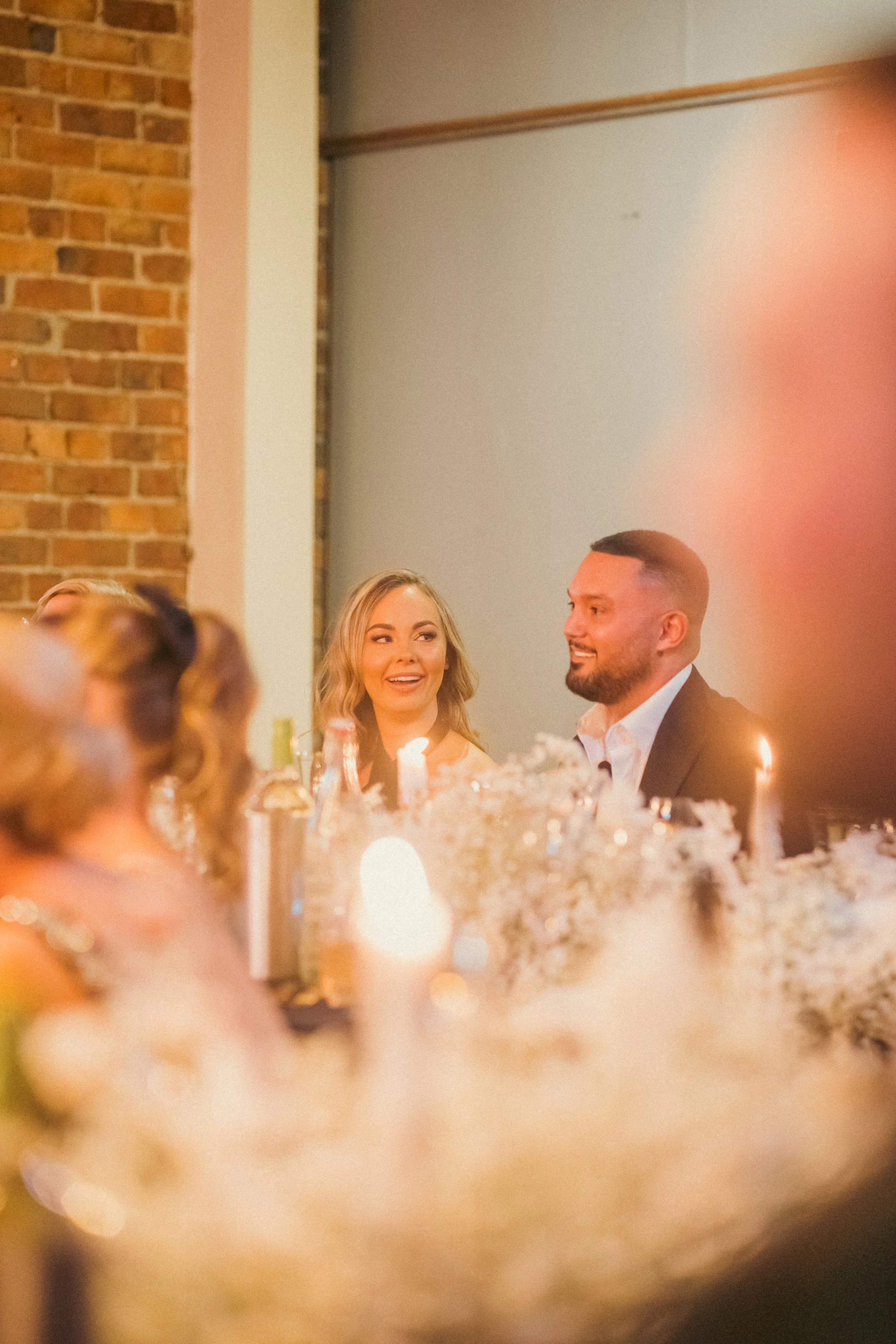 A couple smiling at a wedding reception, sitting at a table with candles and floral centerpieces, with guests and brick wall in the background.