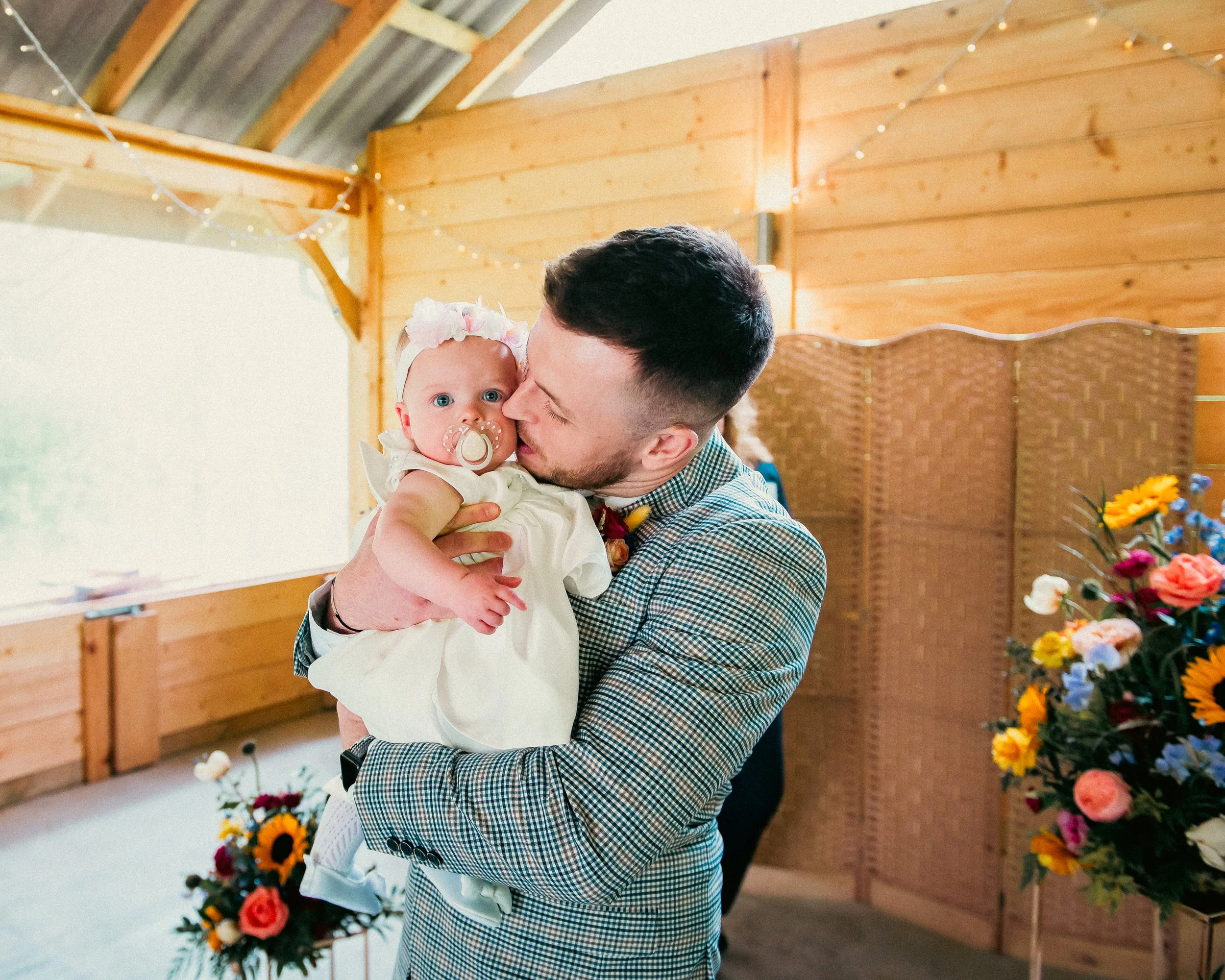 A man holding a baby girl in a cozy, wooden room decorated with flowers and string lights, sharing a tender moment.