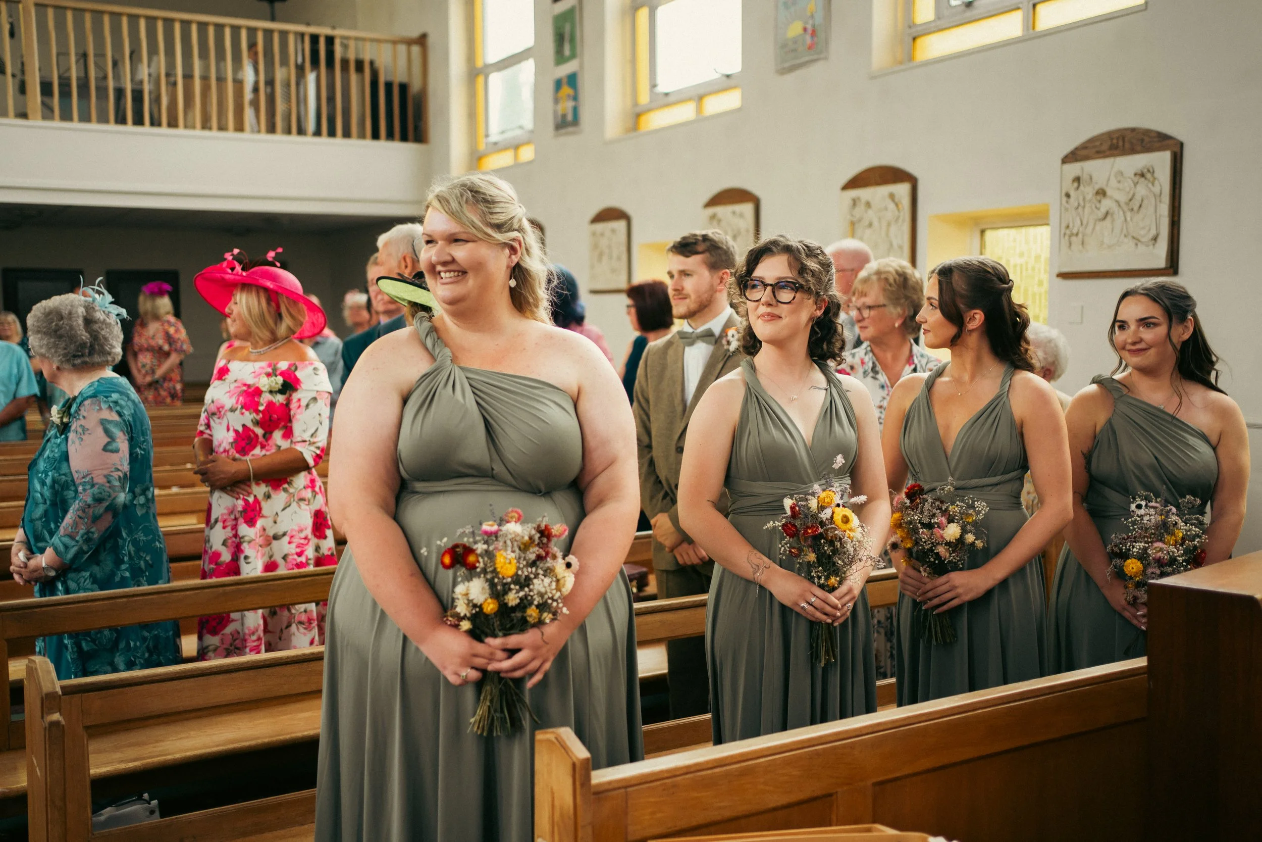 A group of bridesmaids in grey dresses holding bouquets standing in a church during a wedding ceremony.