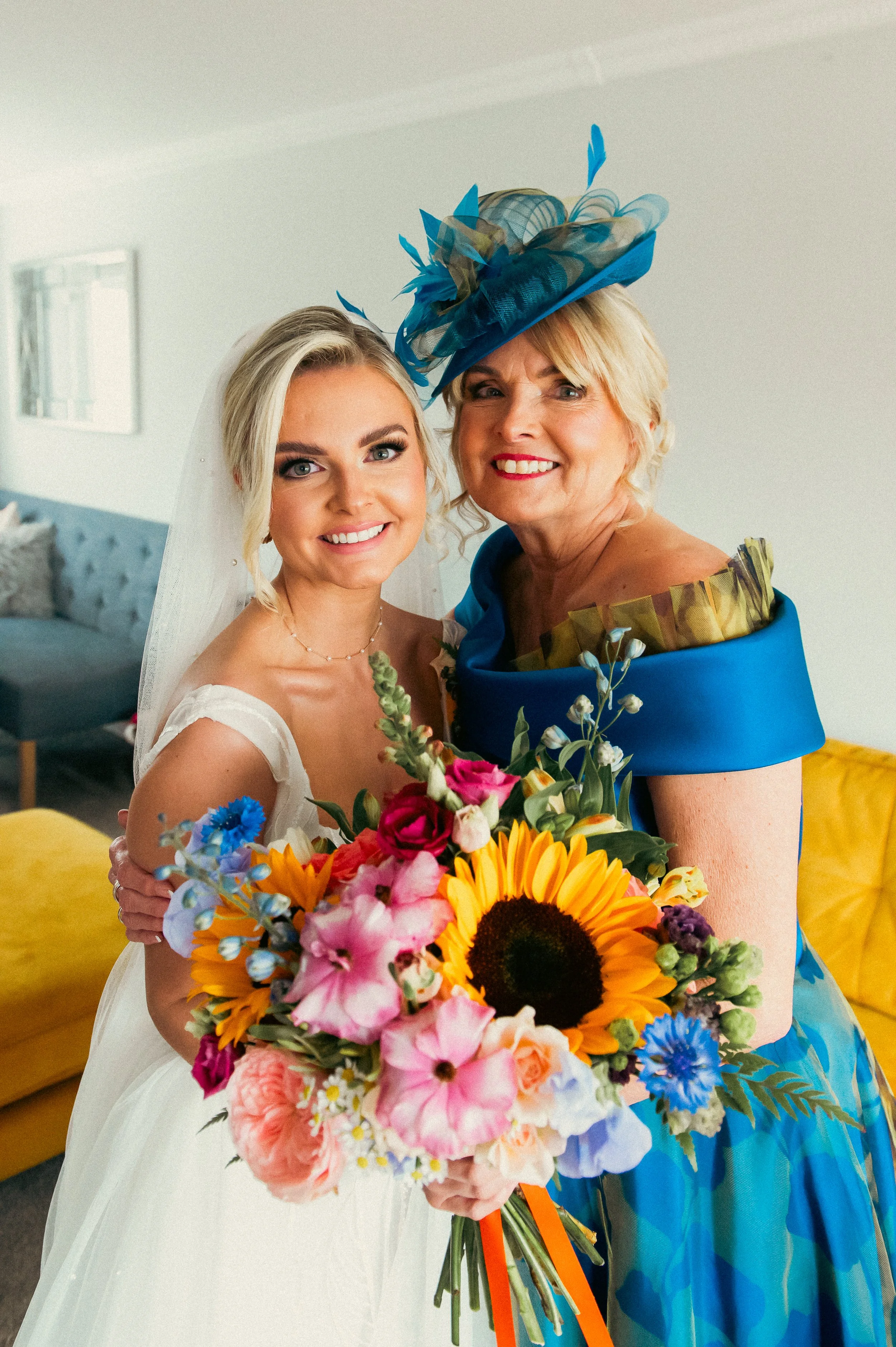 A bride in a white wedding dress smiling and holding a colorful bouquet of flowers, standing next to an older woman in a bright blue dress and matching hat, both smiling and posing for a photo indoors.