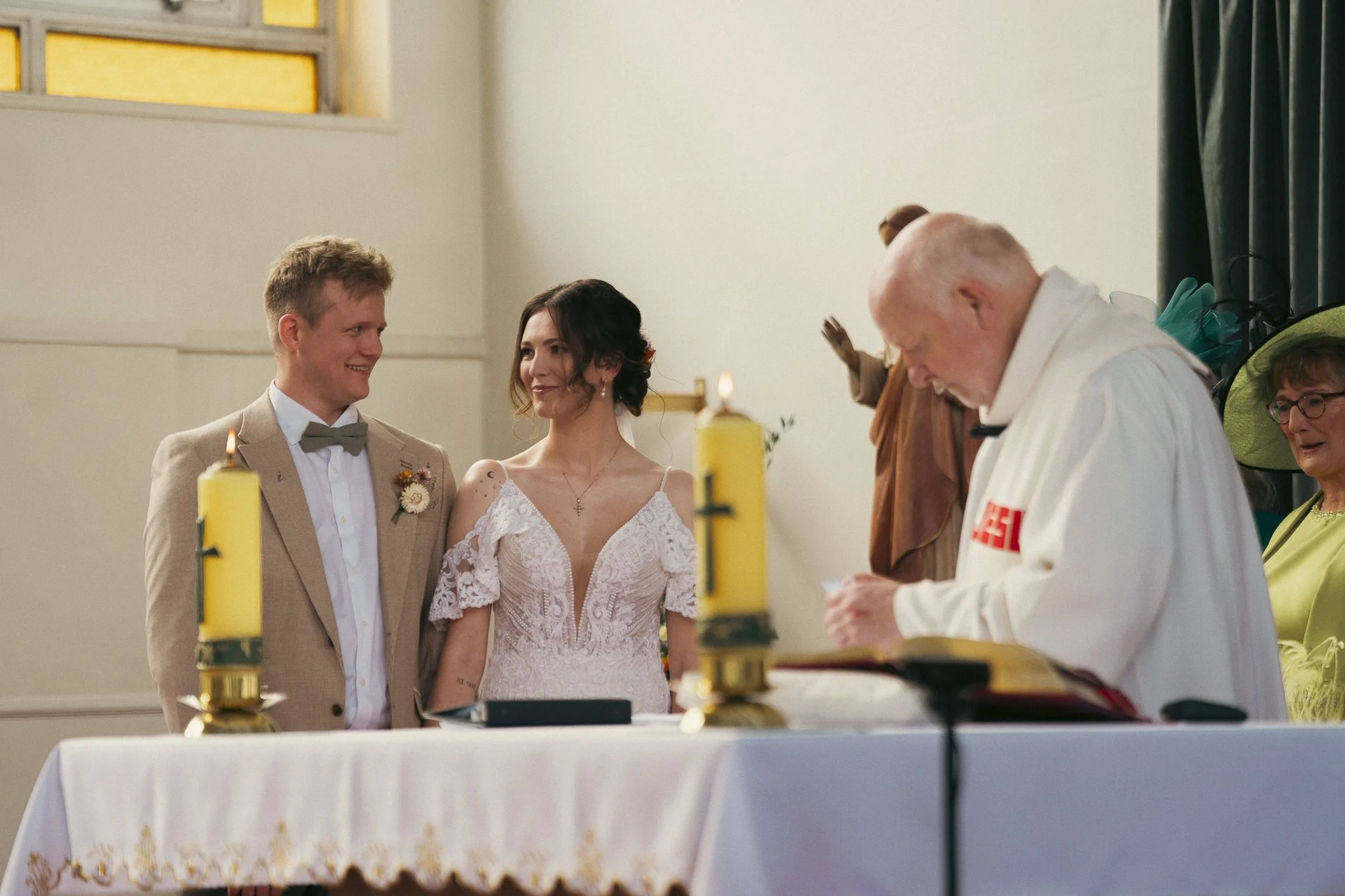 A bride and groom standing at an altar with a priest officiating wedding ceremony, candles on the altar, and guests in the background.
