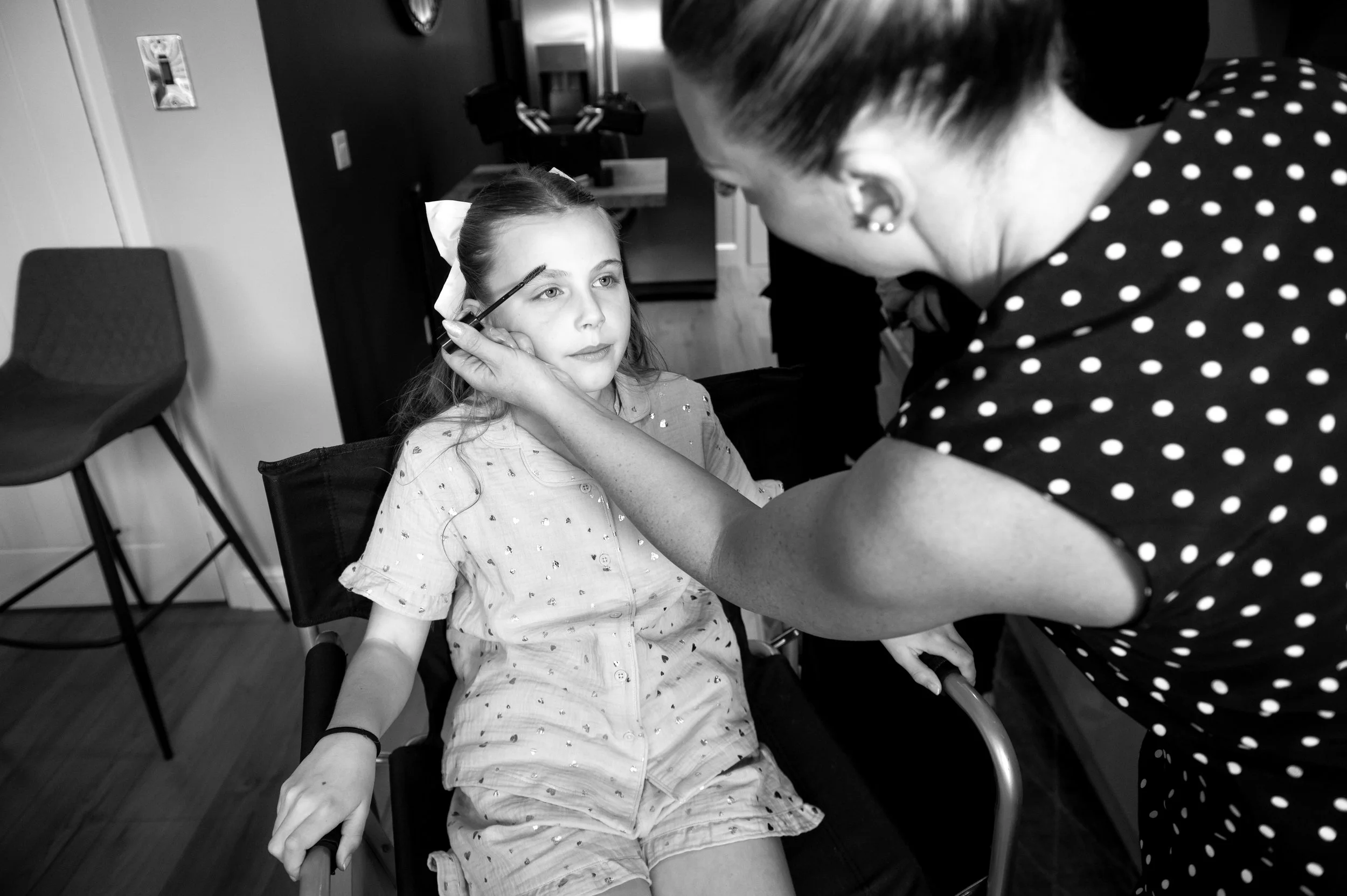 A young girl sitting in a chair, having her eyebrows makeup applied by a woman in a polka dot shirt, inside a room with chairs and a counter in the background.