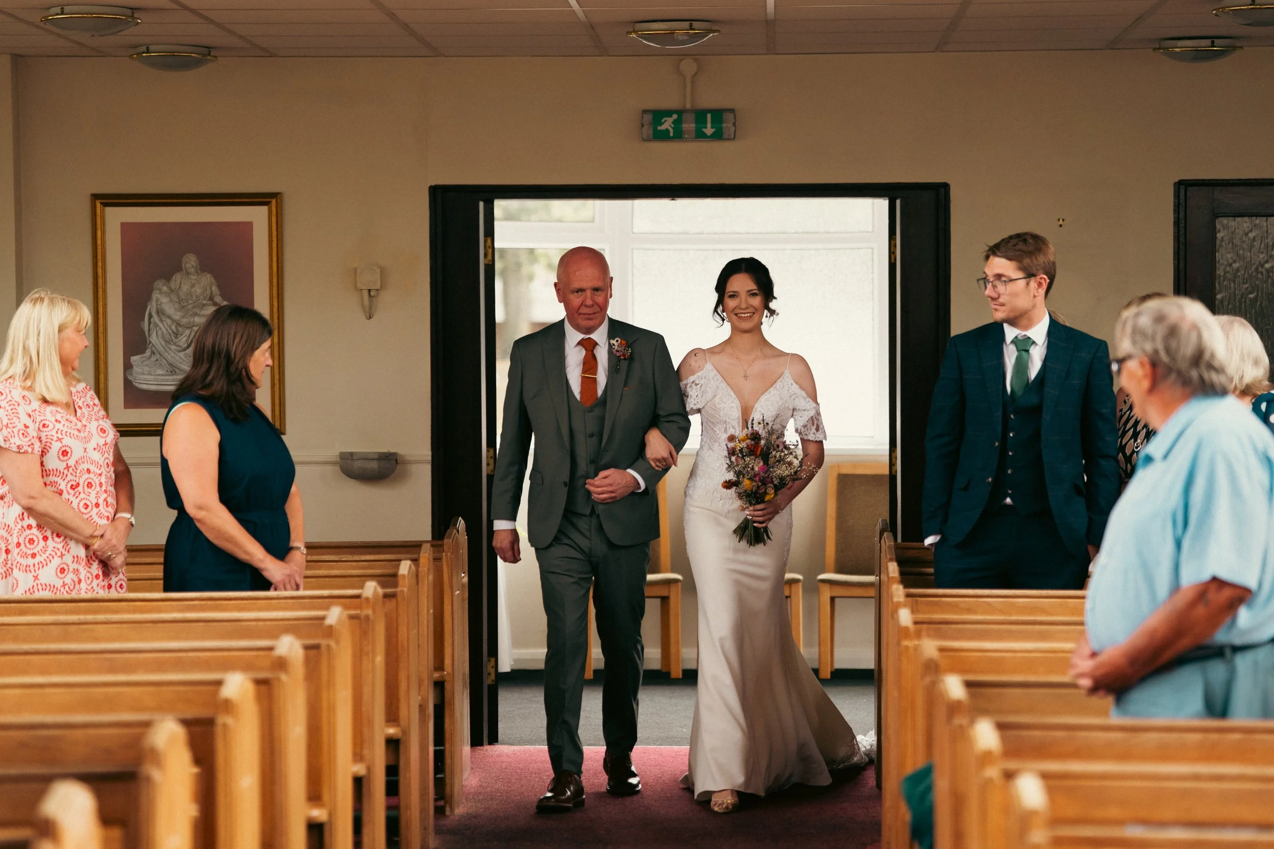A bride with a bouquet of flowers walking down the aisle into a church, escorted by an older man in a suit. Several guests are standing and looking at her as she enters.