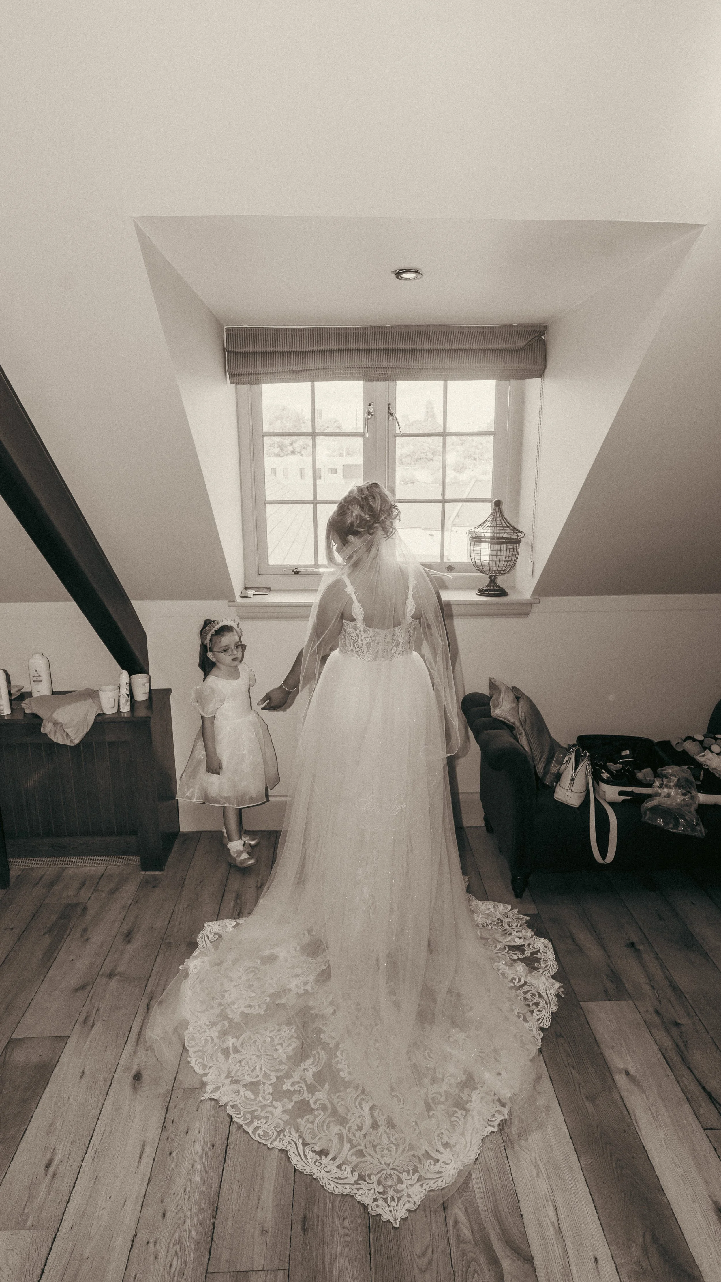 A bride in a wedding gown with a veil holding hands with a young girl in a white dress, standing in front of a window in a room with wooden flooring.