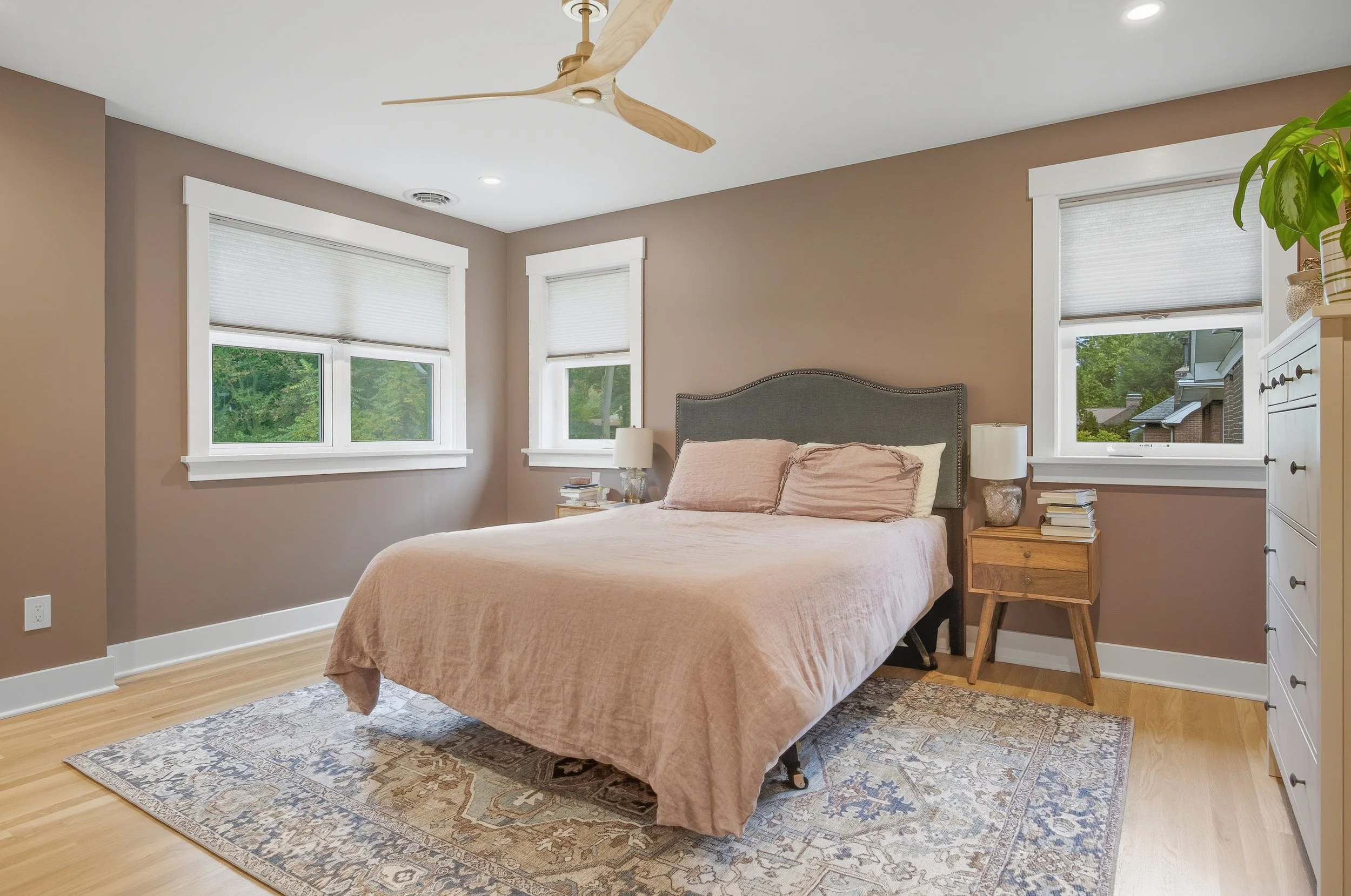 A bedroom with a beige bedspread, two beige pillows, and a gray upholstered headboard. There are two nightstands with books and lamps, three windows with white blinds, a wooden floor, a patterned area rug, a white dresser, and a ceiling fan.