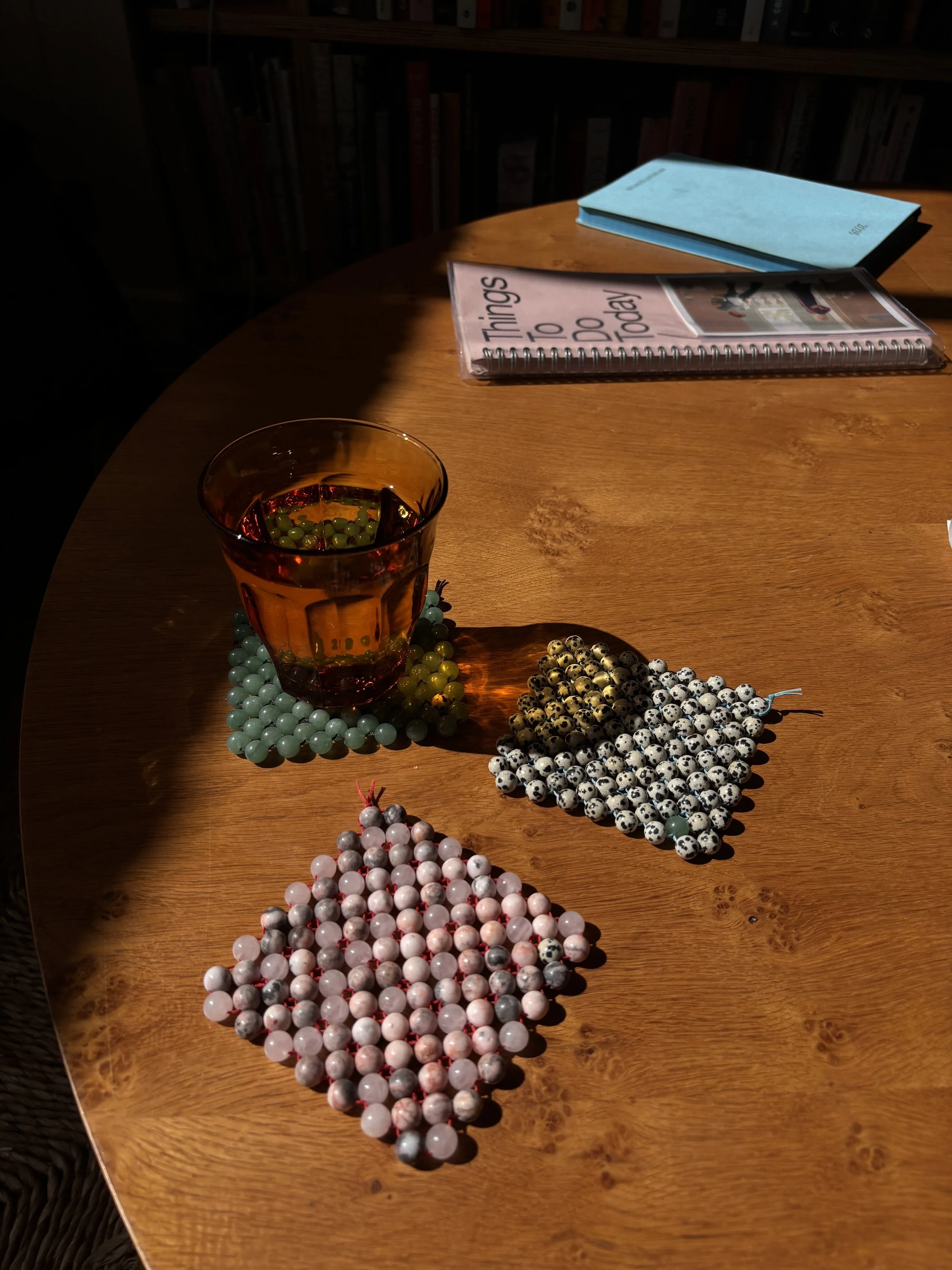 A wooden table with a glass of red beverage, surrounded by various necklaces made of beads in colors including pink, white, black, and teal. In the background, there are notebooks, one pink with spiral binding and the other blue, on the table.