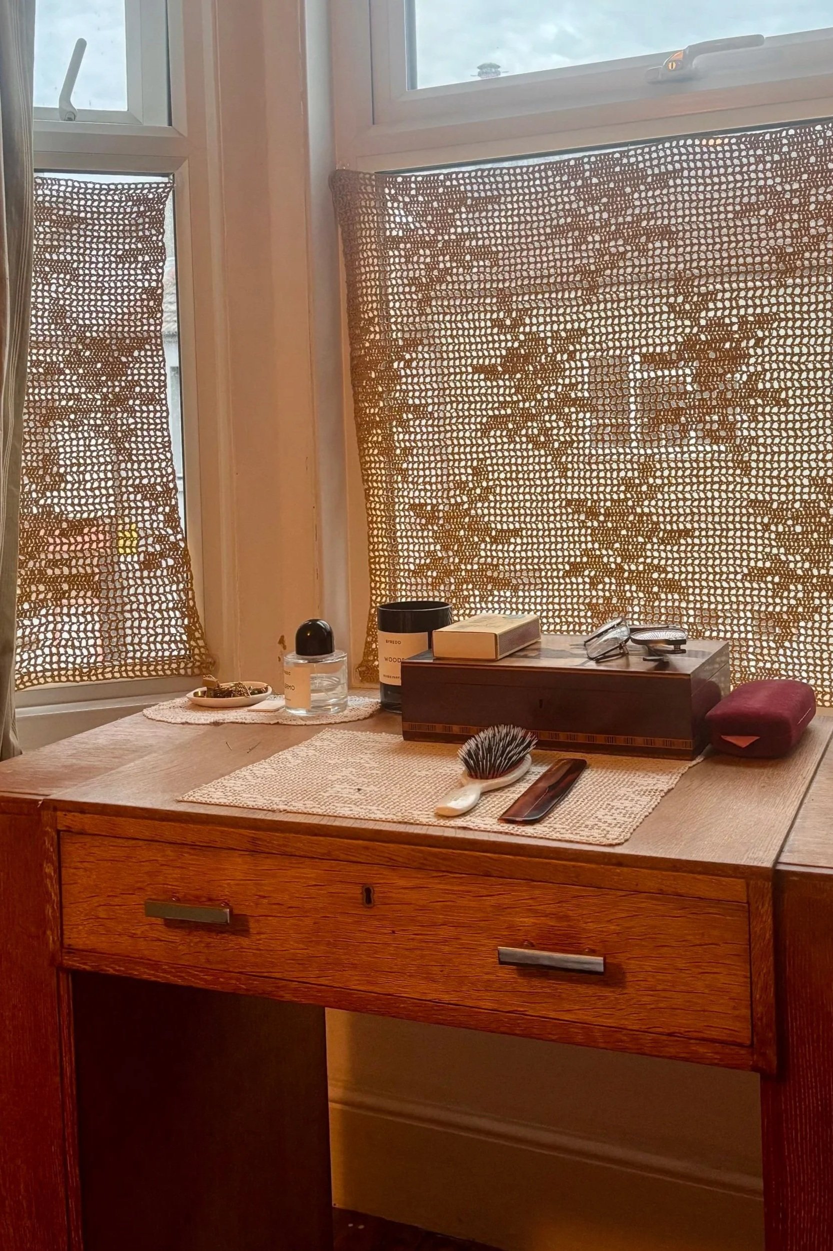 A wooden dresser with a woven placemat on top, holding a hairbrush, a smartphone, and other small items. Behind it are two windows with netted curtains and a small wooden box. Various personal items and a pair of glasses rest on the window ledge.