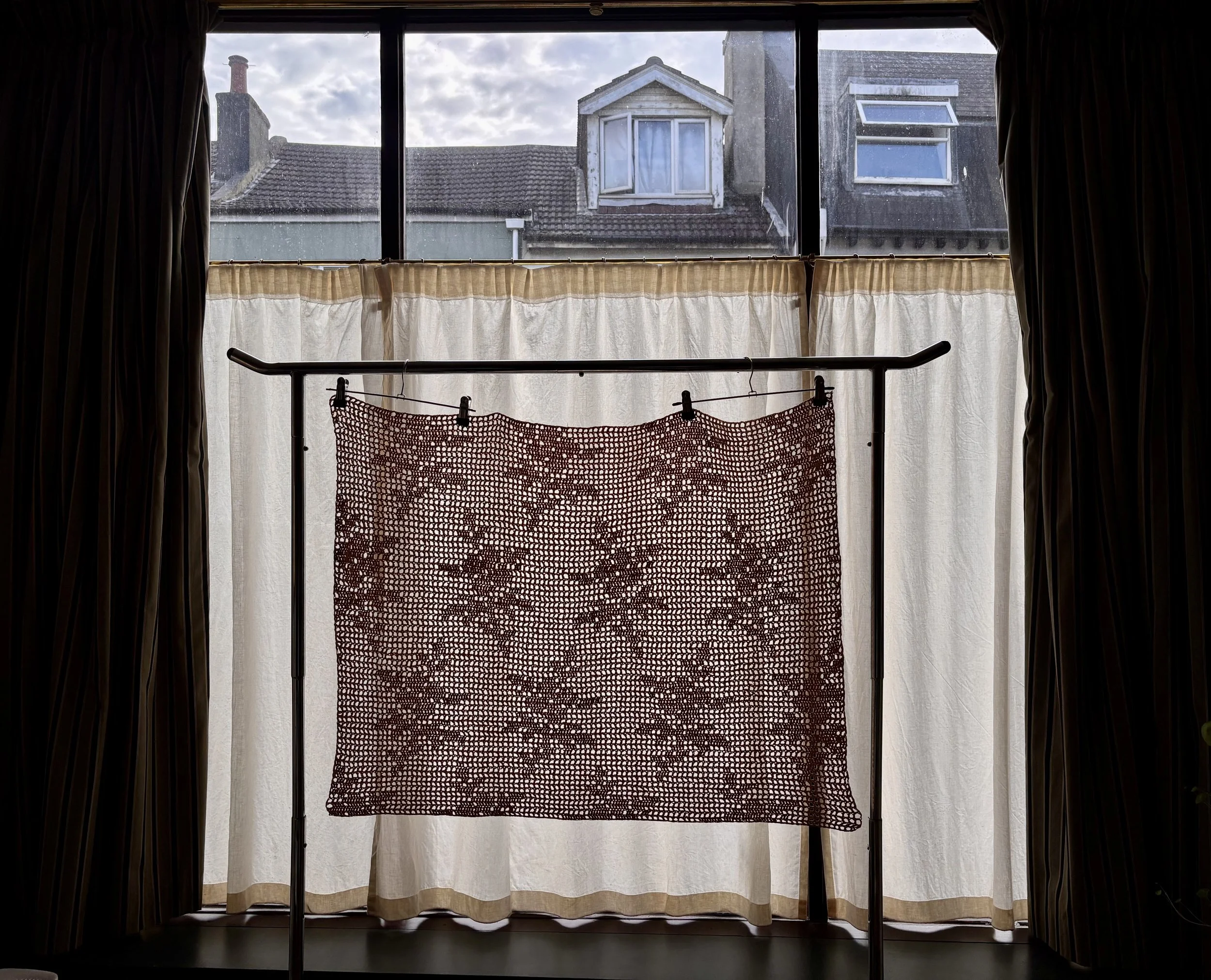 View of neighboring rooftops, buildings with skylights, through a large window with curtains, a clothes drying rack with a red netted cloth inside