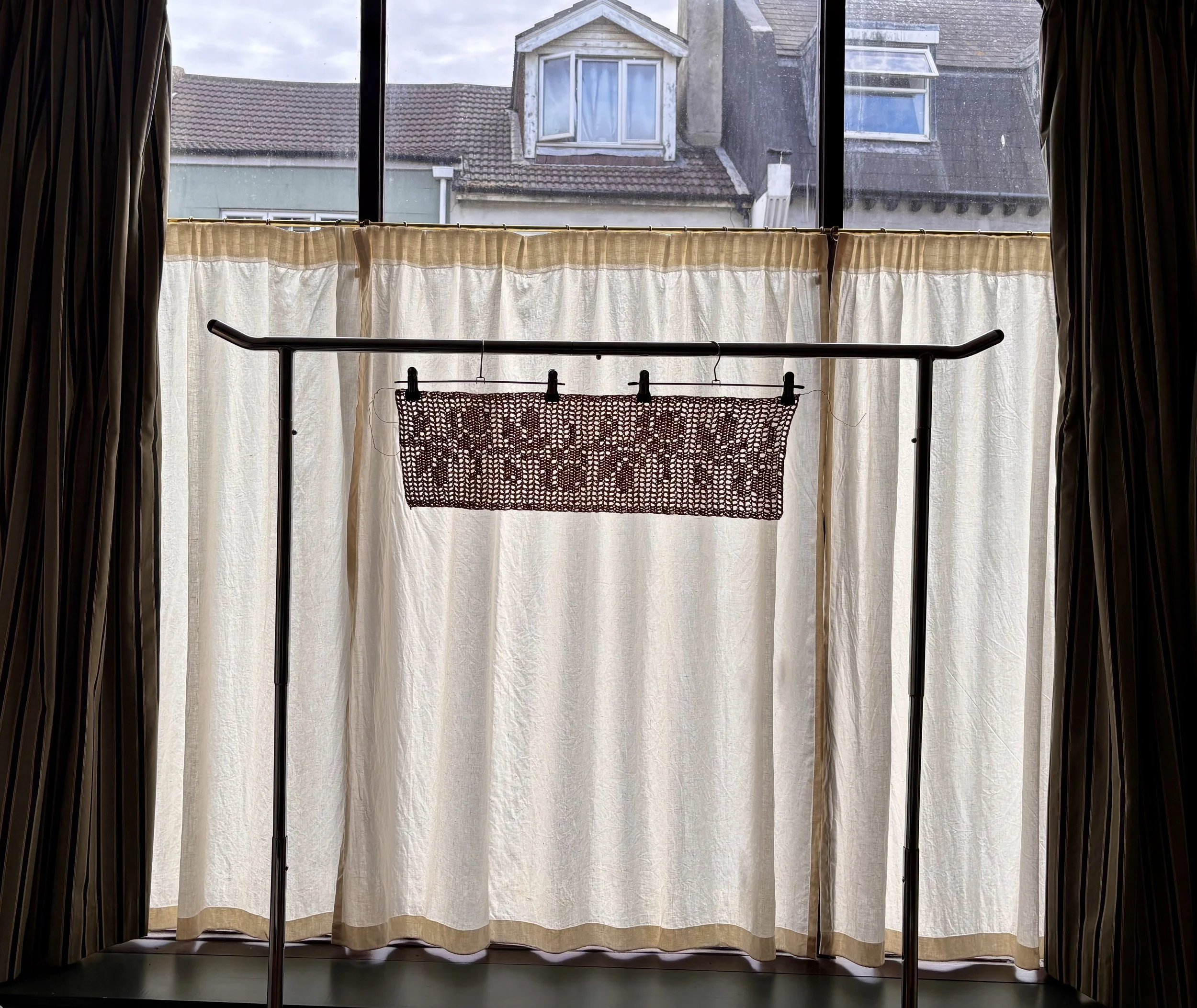 Indoor view through a large window with beige curtains, showing neighboring rooftops and a cloudy sky outside.