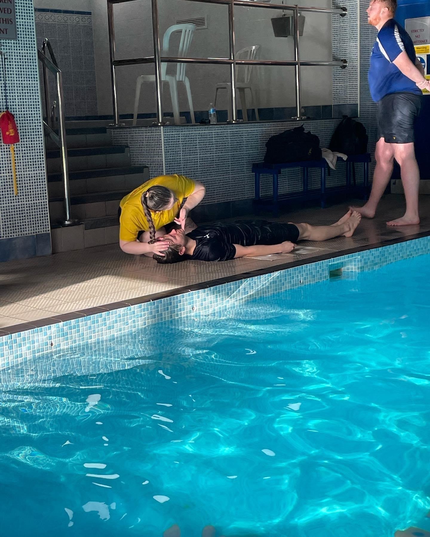 A person in a black shirt lies on the ground next to a swimming pool, receiving aid from a person in a yellow shirt. Another person in a blue shirt stands nearby, observing the scene. The setting is an indoor pool area with chairs and bags in the bac
